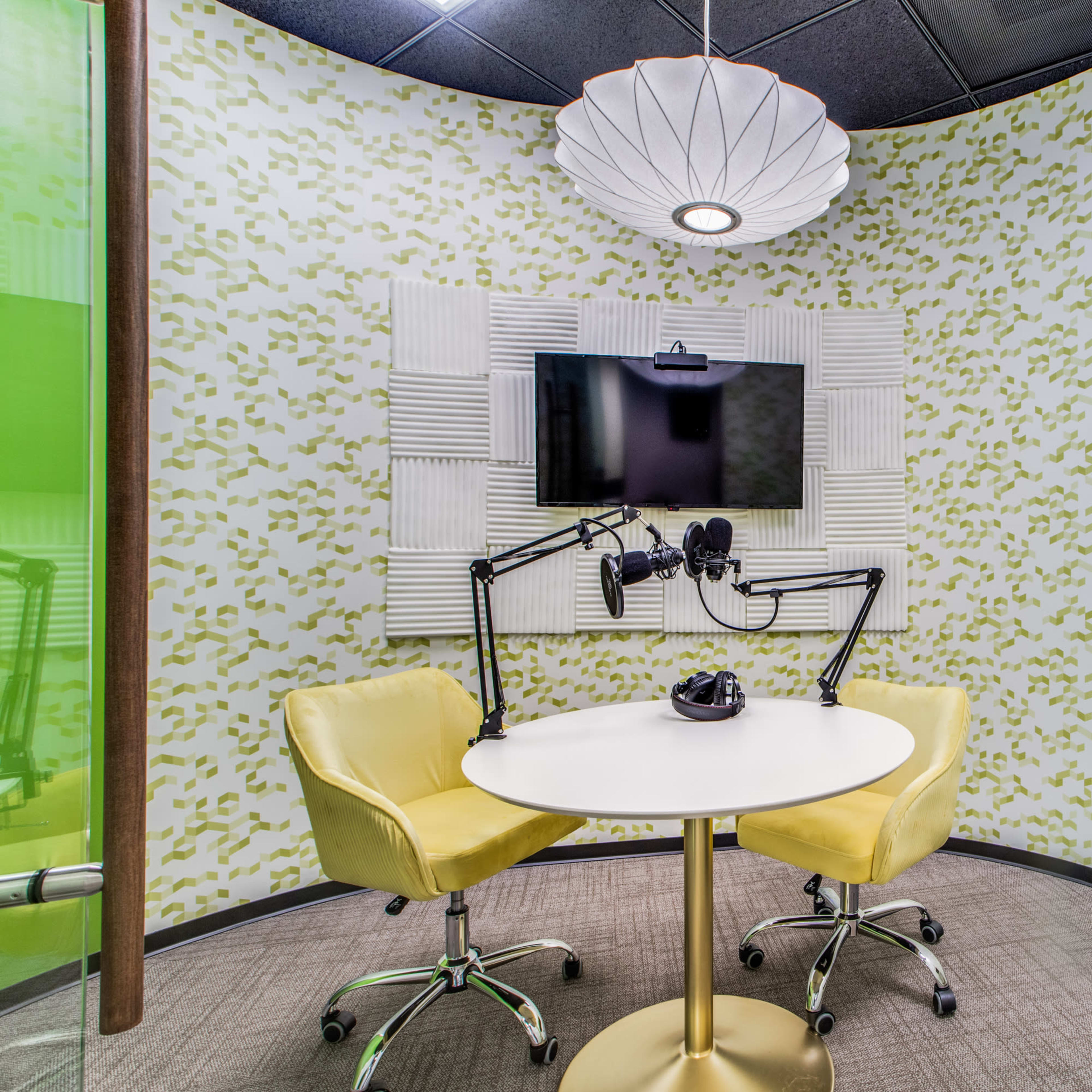 A small recording studio featuring a round table with two yellow chairs, a wall-mounted television, and microphone setups on arms, with a ring light and a decorative wall behind.