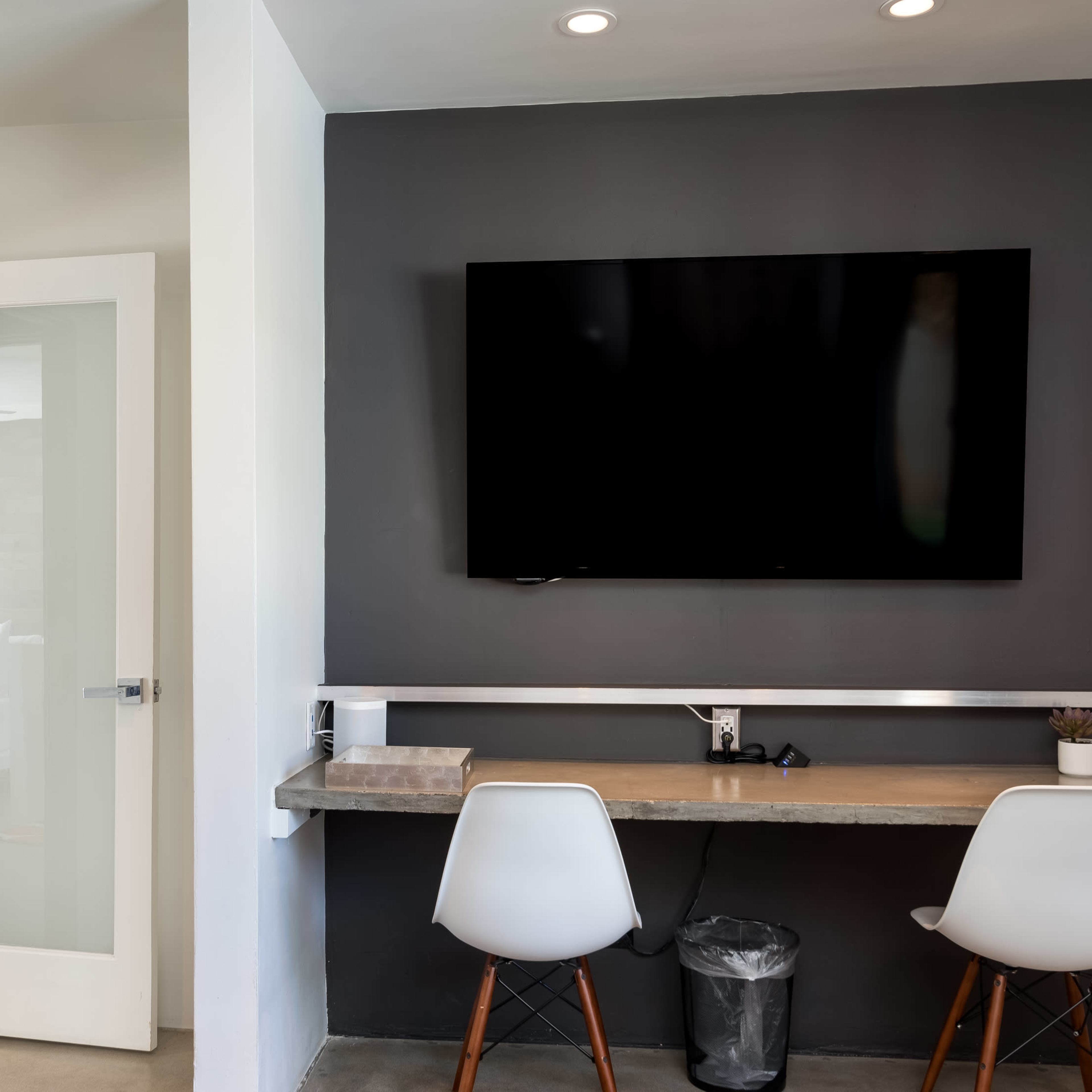 A minimalist workspace features a wall-mounted TV above a simple desk with two chairs and a visible trash can.