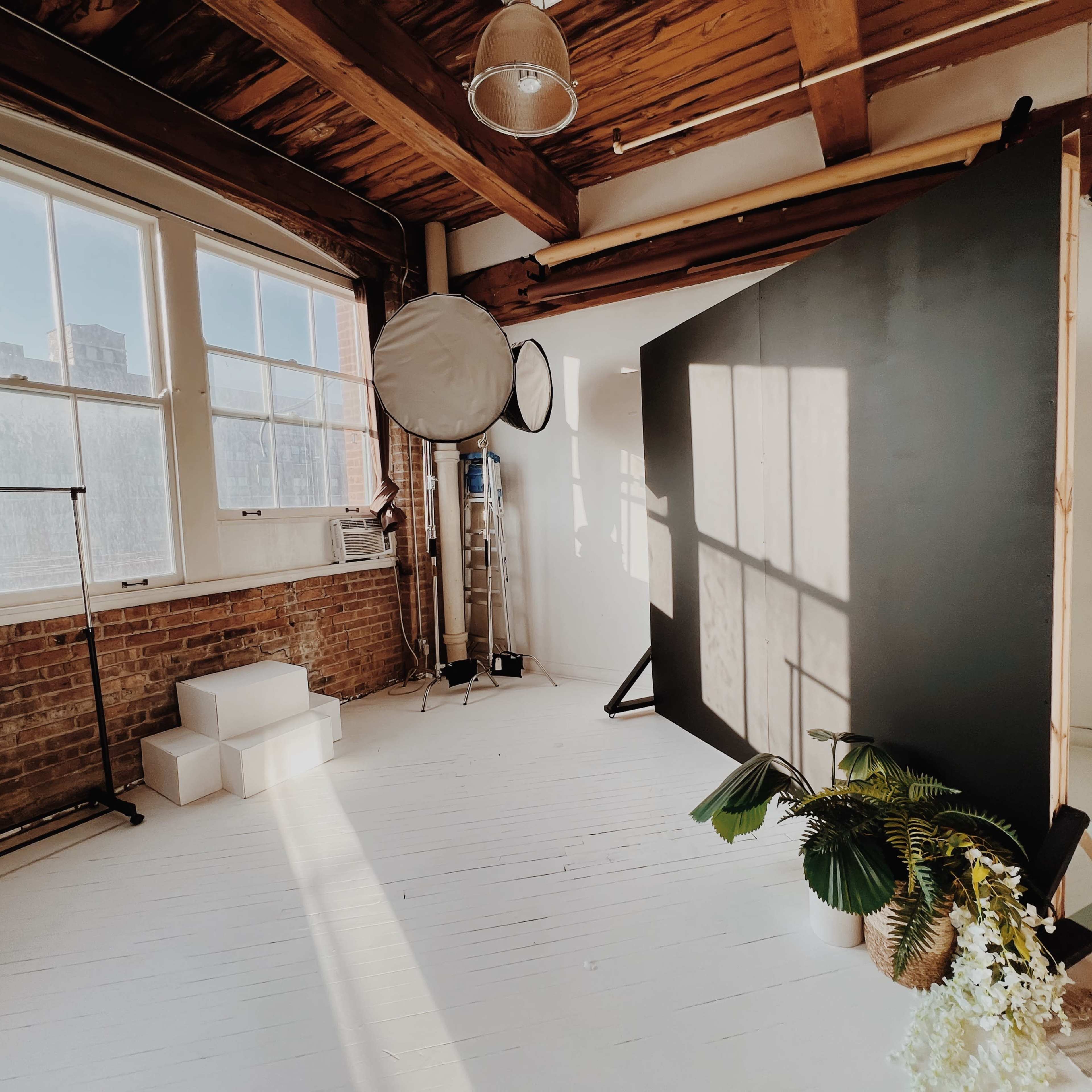A spacious, brightly lit studio with wooden beams, a large window, a dark backdrop, and a hammock chair adorned with plants.