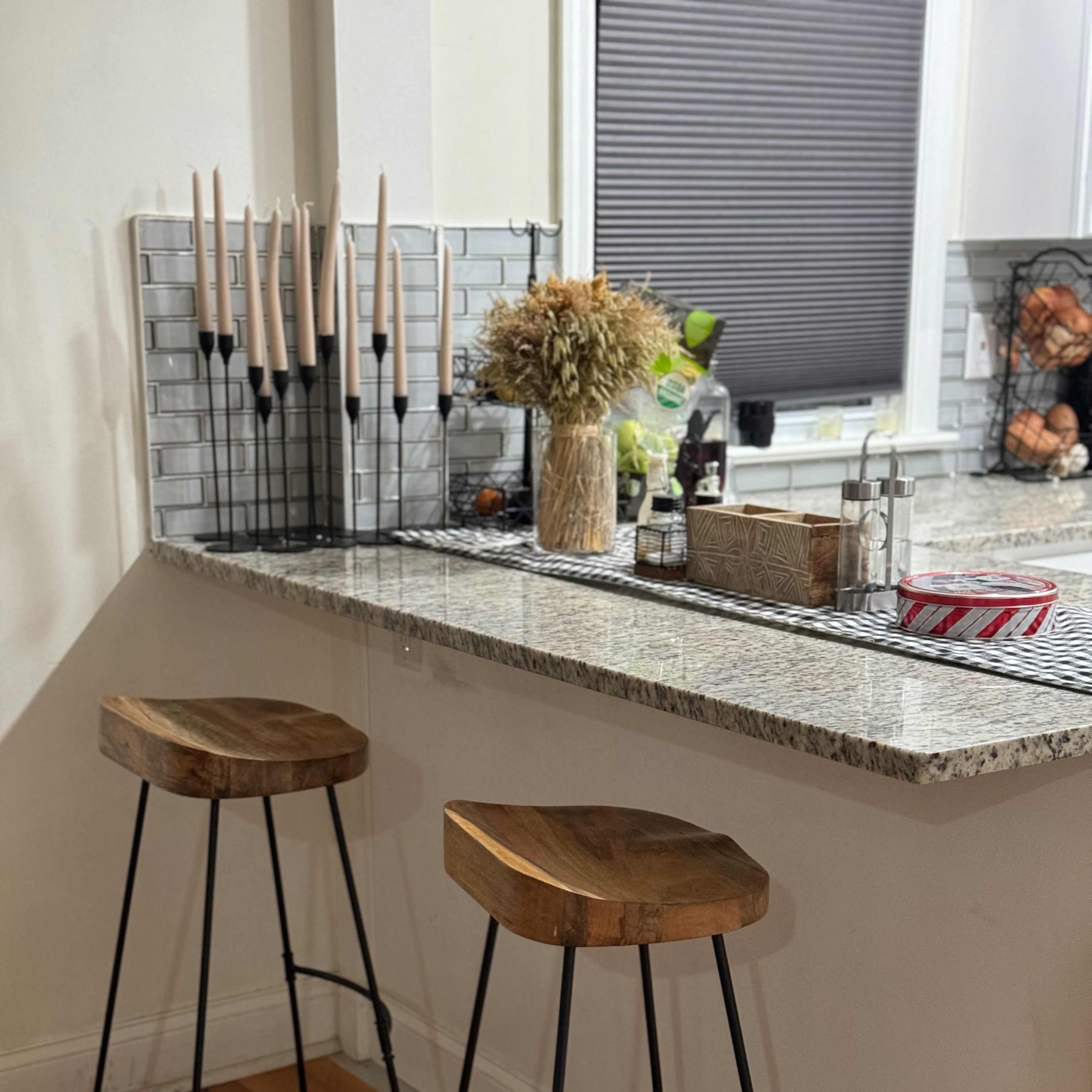 A kitchen bar area with two wooden stools, a granite countertop, and a decorative arrangement of candles and dried flowers in a glass vase.