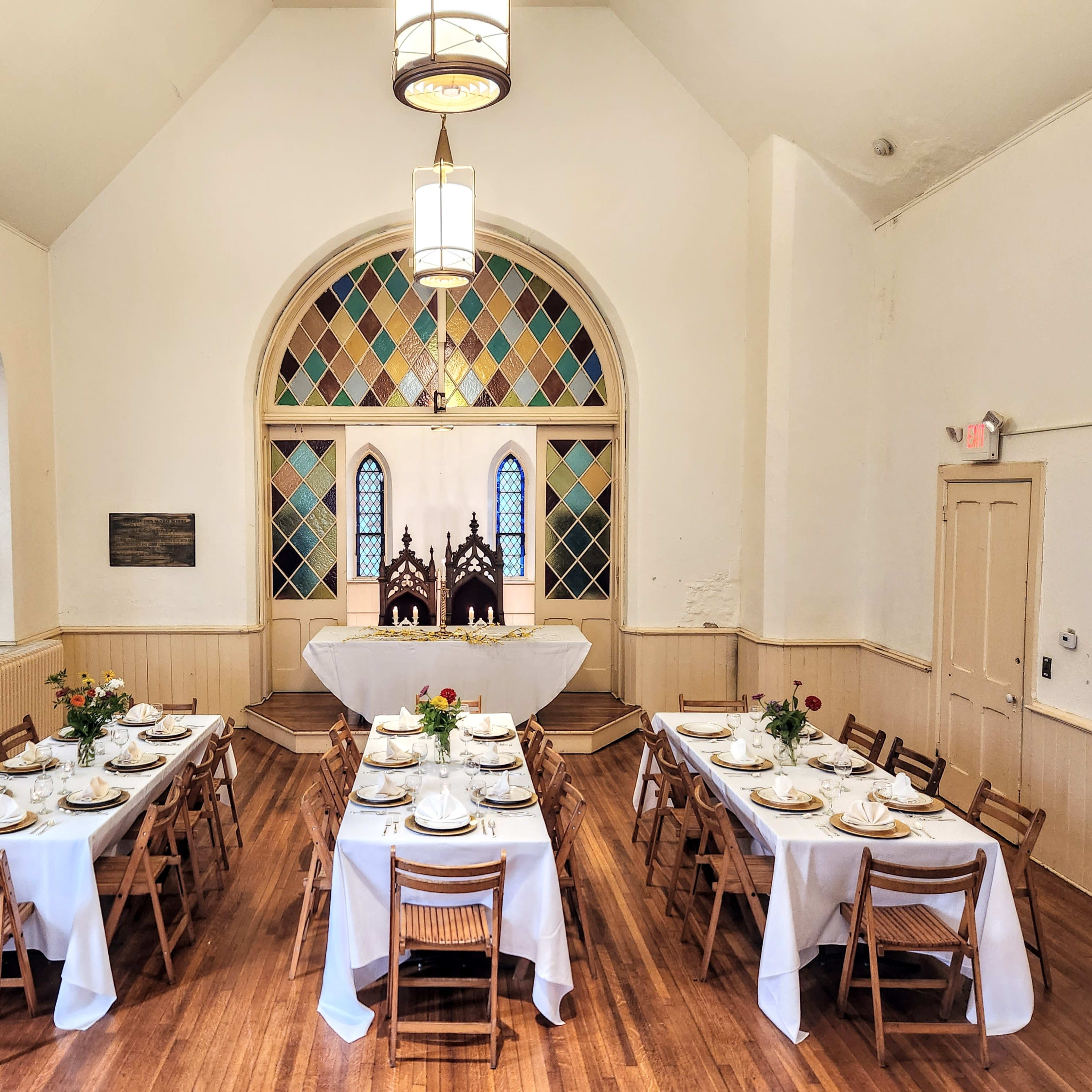 A dining hall features two long tables set with plates and flowers, arranged symmetrically beneath stained glass windows.