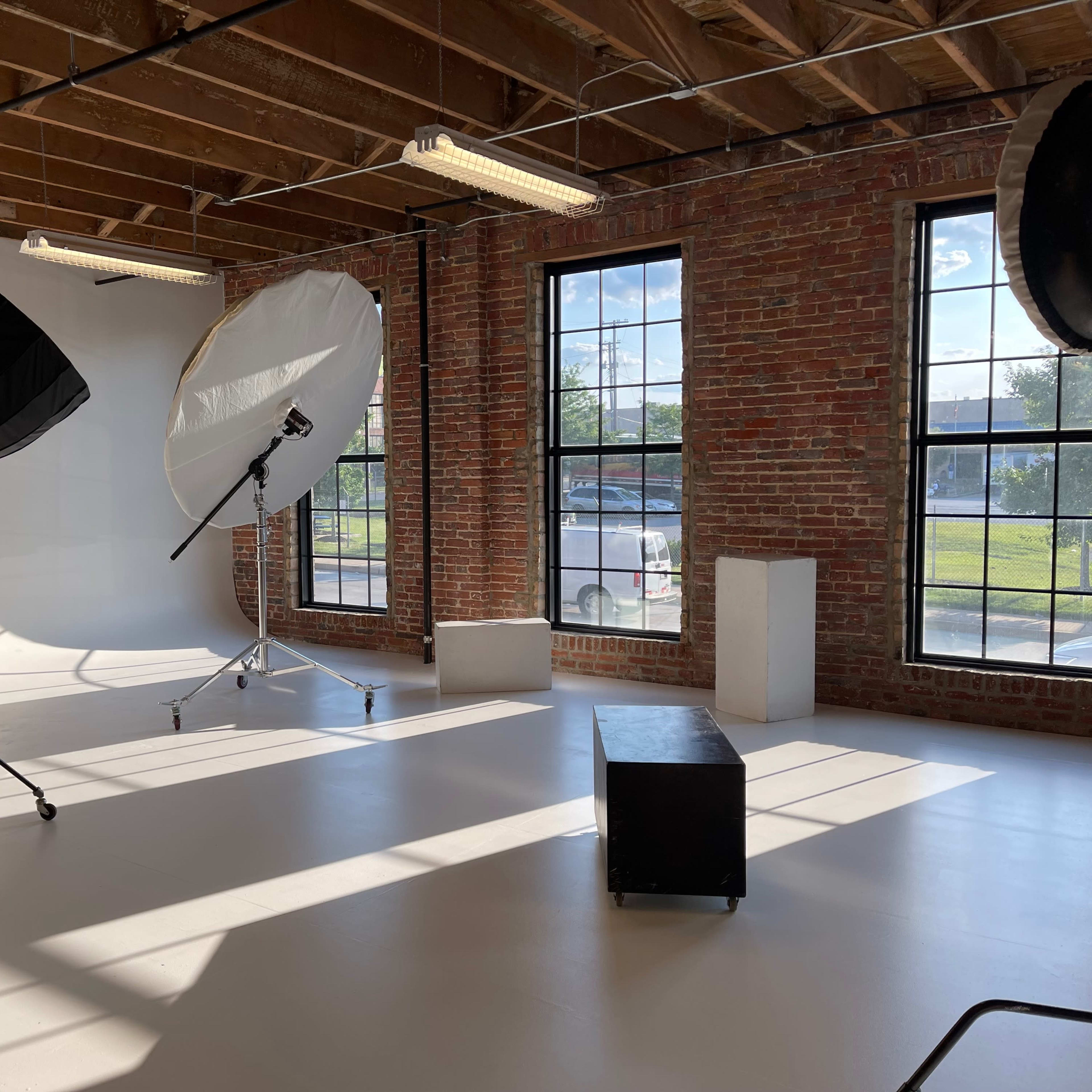 A spacious studio with large windows, brick walls, and photography lighting equipment set up on a white floor.