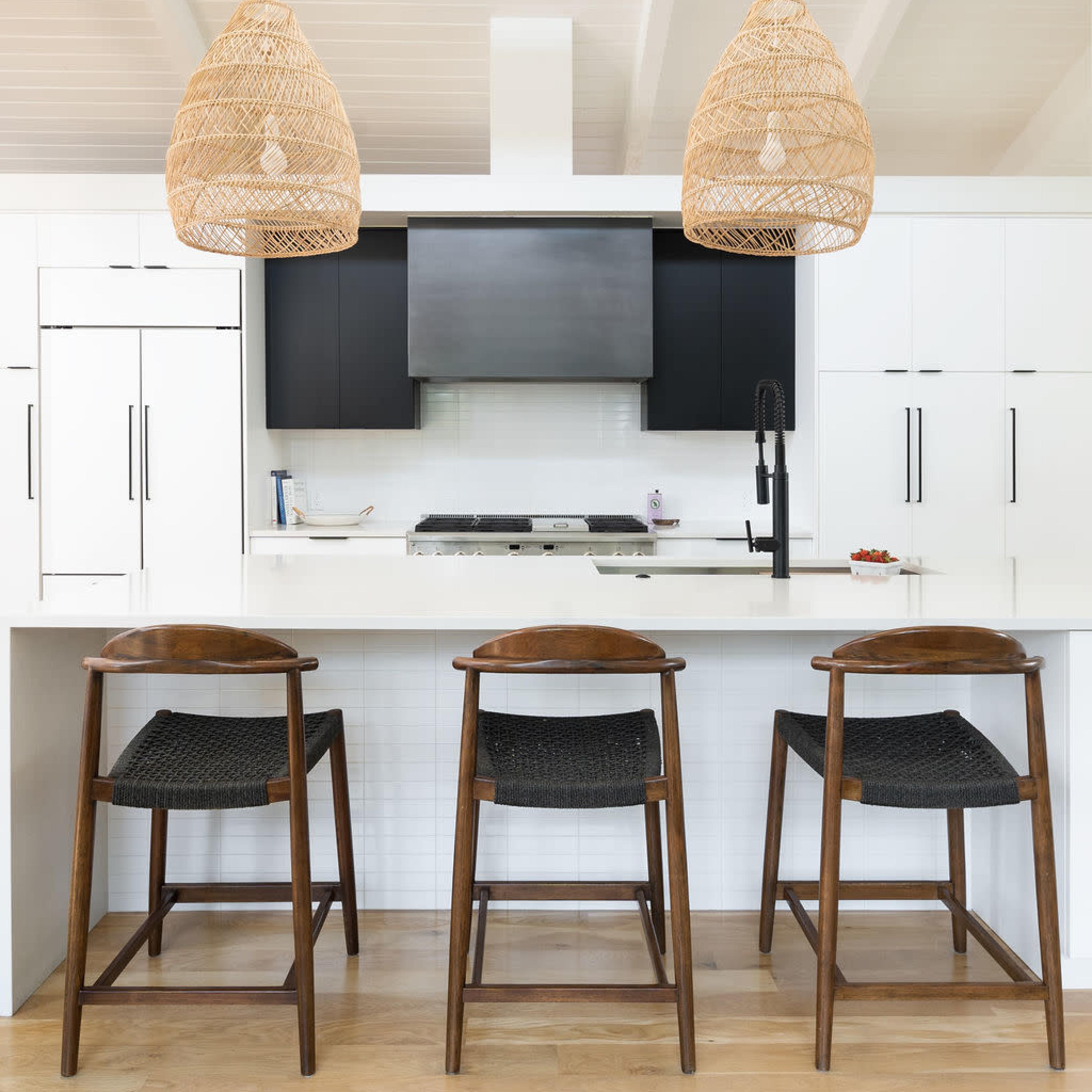 A modern kitchen features a central island with three wooden stools, white cabinetry, and two woven pendant lights hanging overhead.