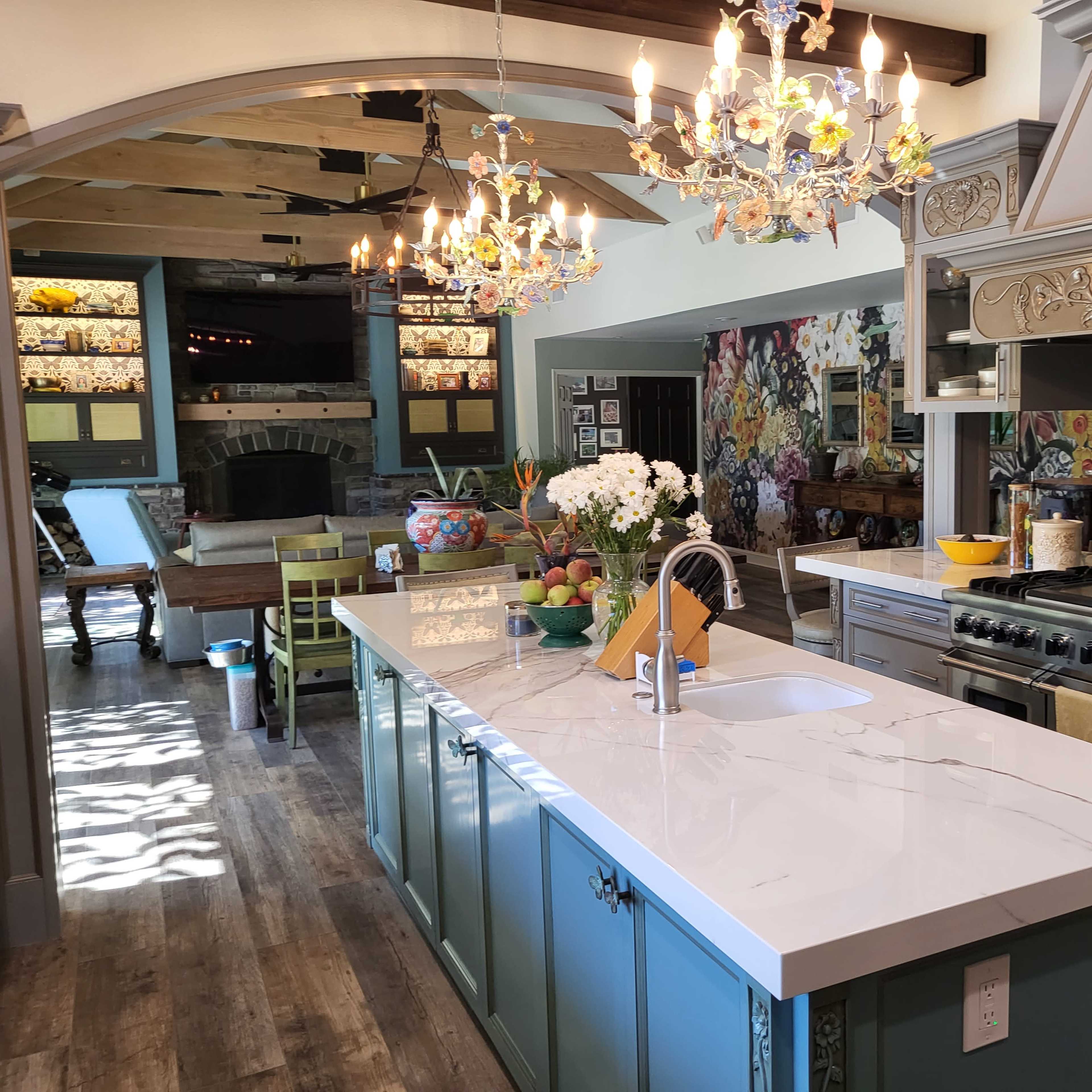 The kitchen features a large island with a white countertop, blue cabinetry, and ornate chandeliers, surrounded by floral wallpaper and wood accents.