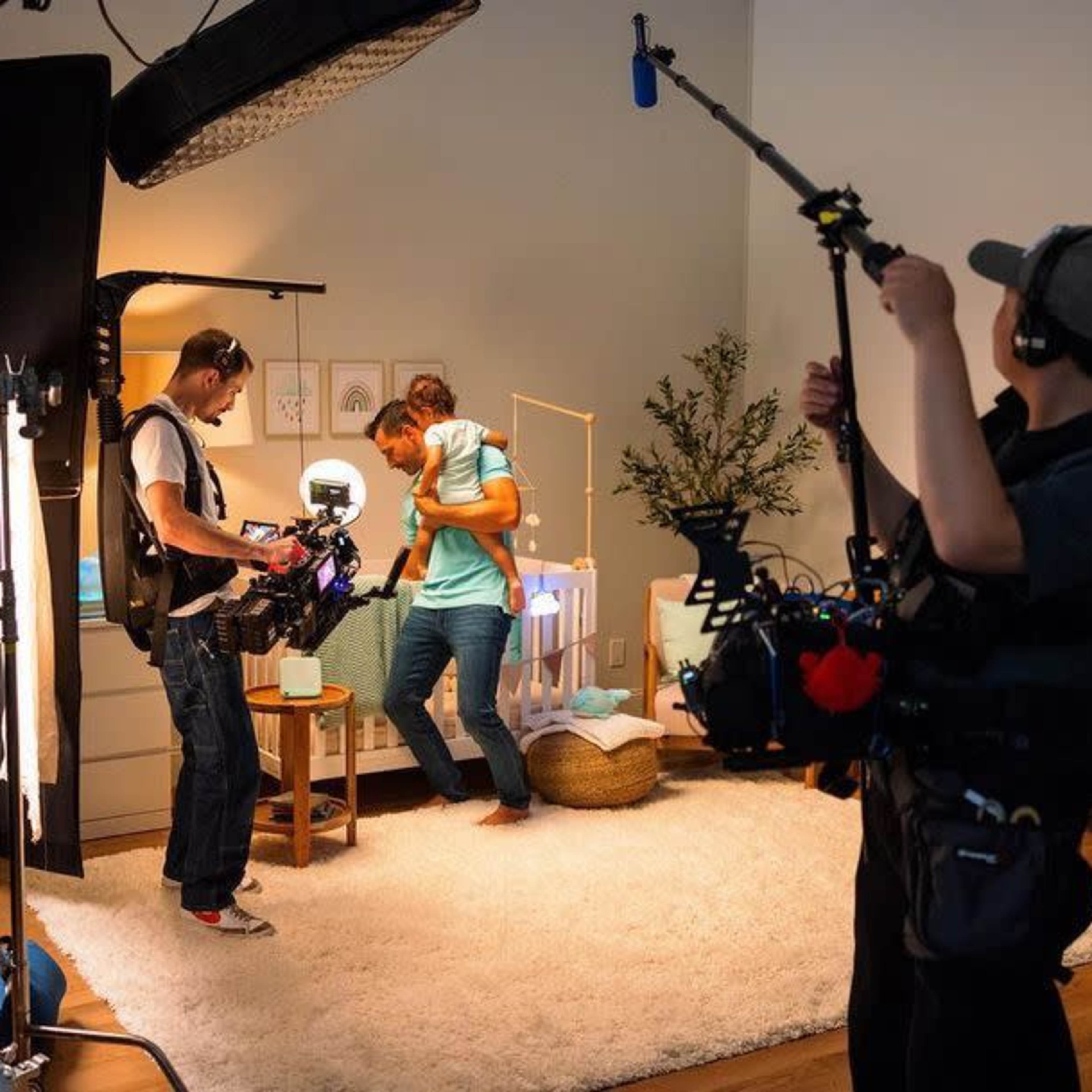 A film crew sets up equipment in a room while a person interacts with another in a cozy living space.