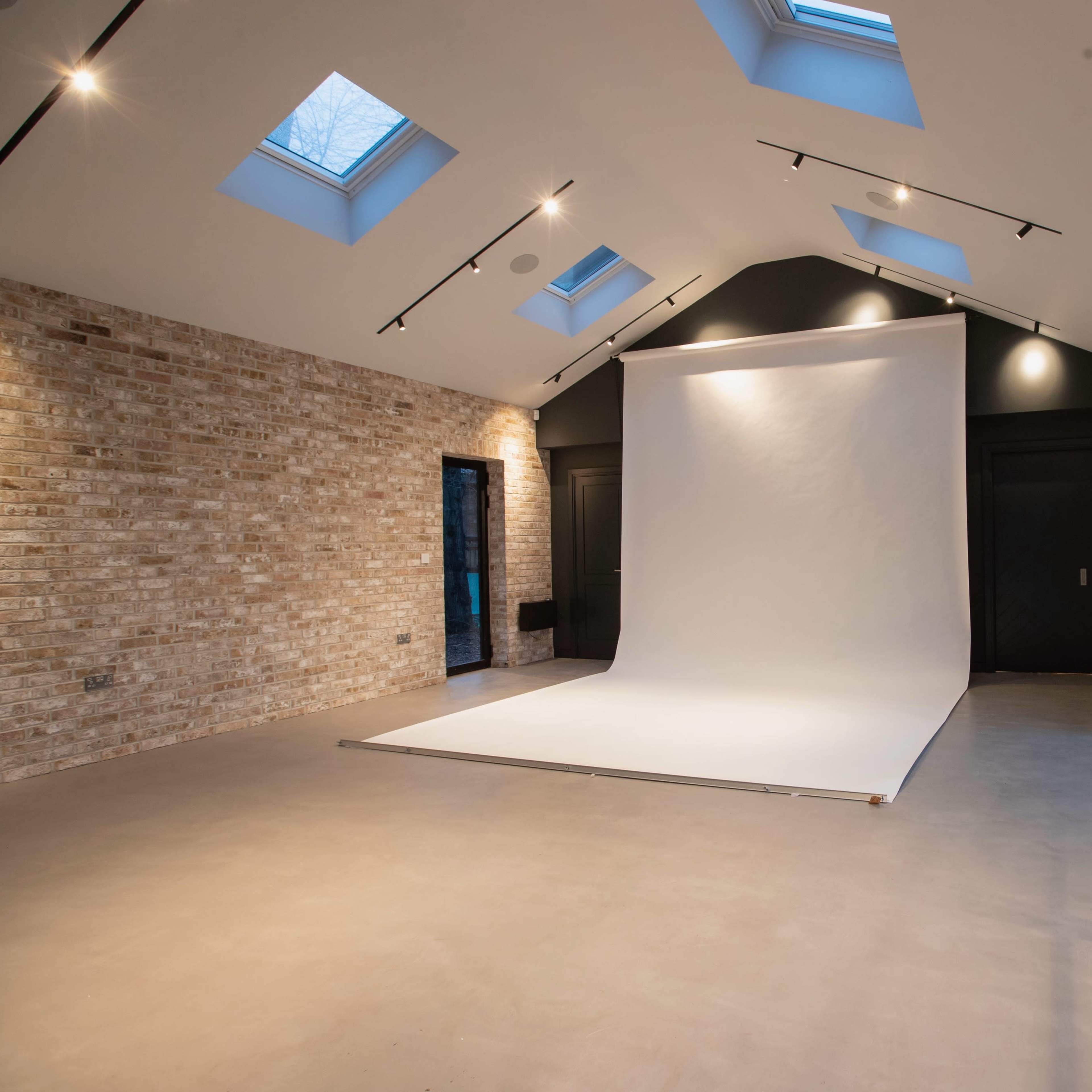 A spacious studio with brick walls, a large white backdrop, and skylights illuminating the area.