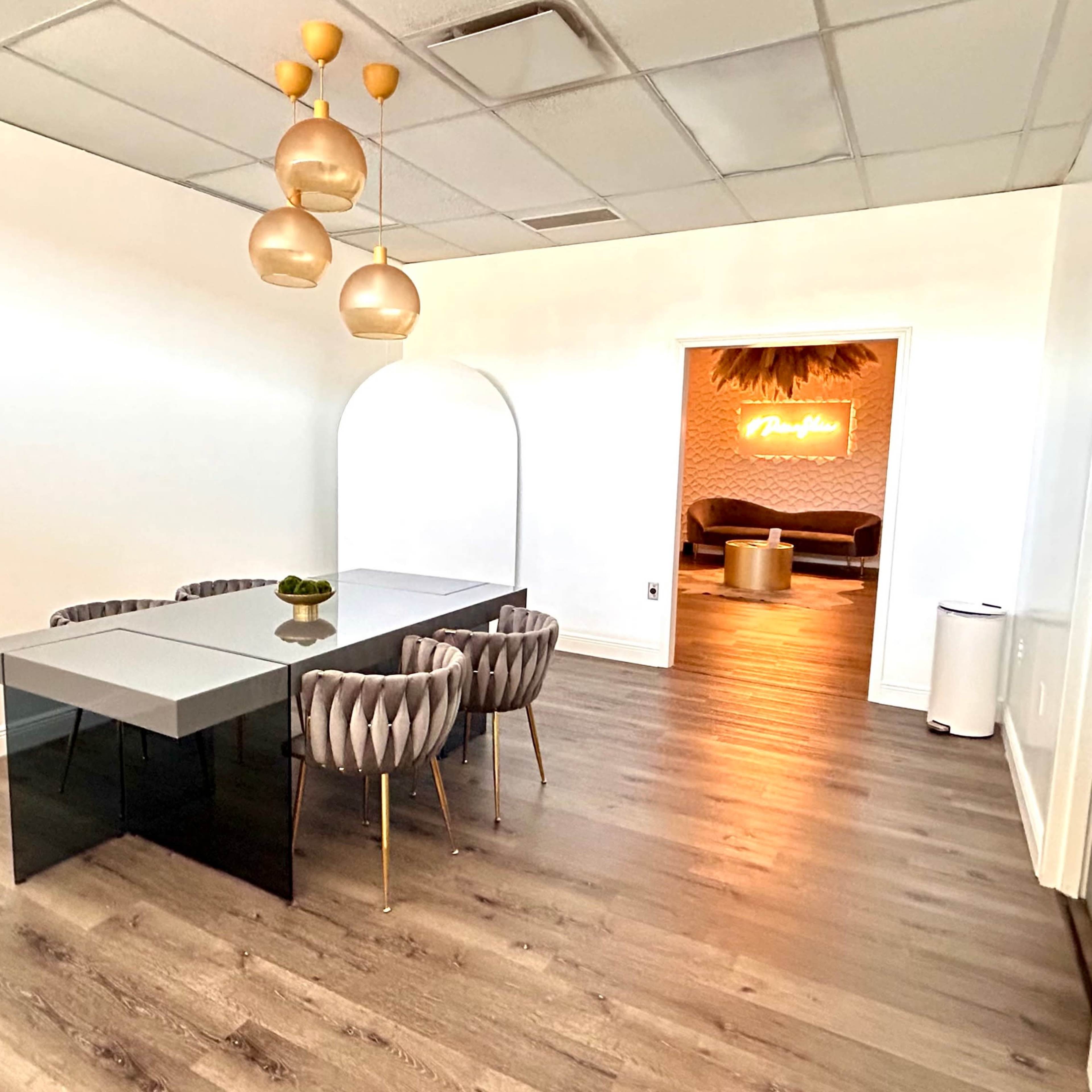 A modern conference room features a sleek glass table surrounded by tufted chairs, with a doorway leading to a lounge area illuminated by neon signage.