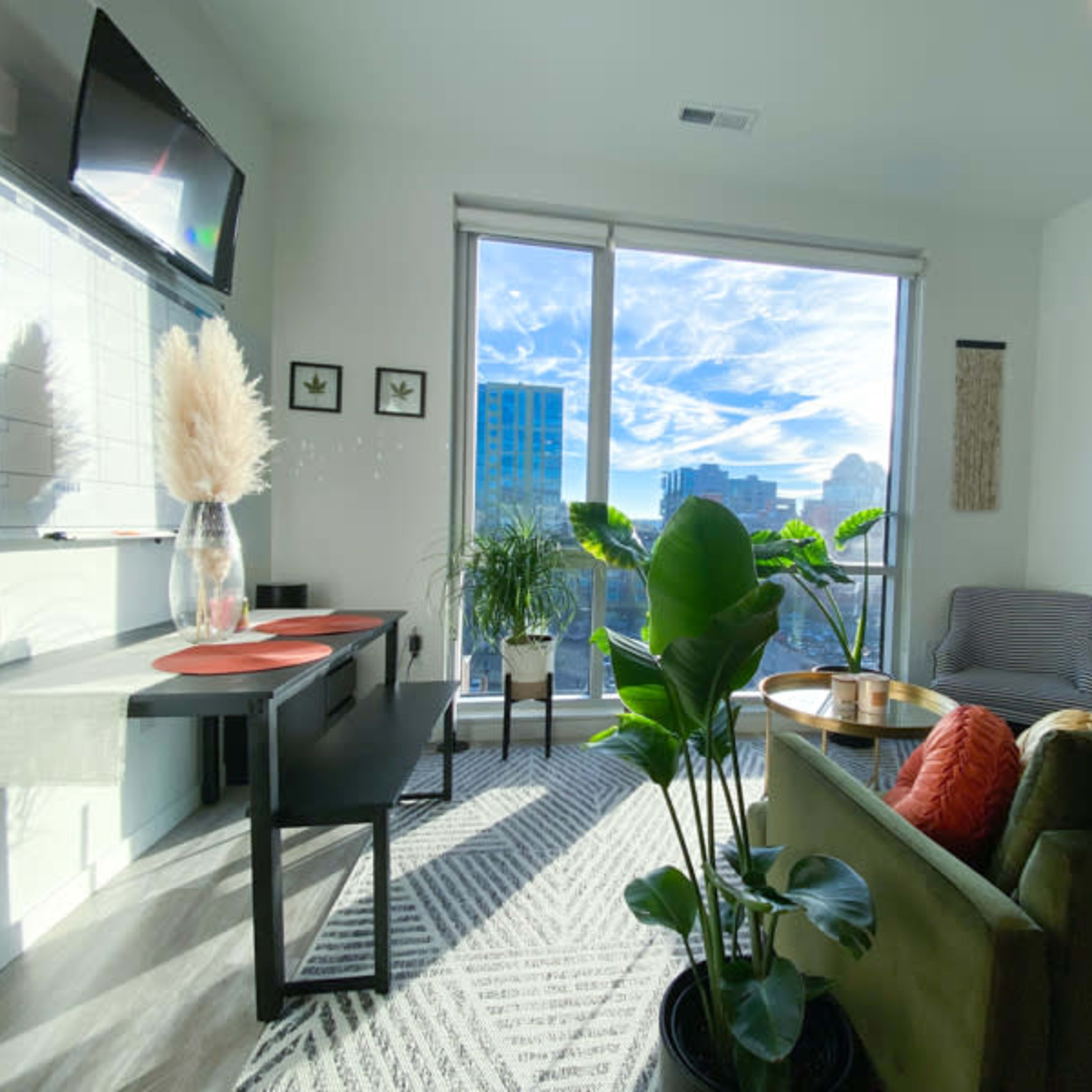 A modern living room features a green sofa, a dining table with chairs, and large windows offering a city skyline view.