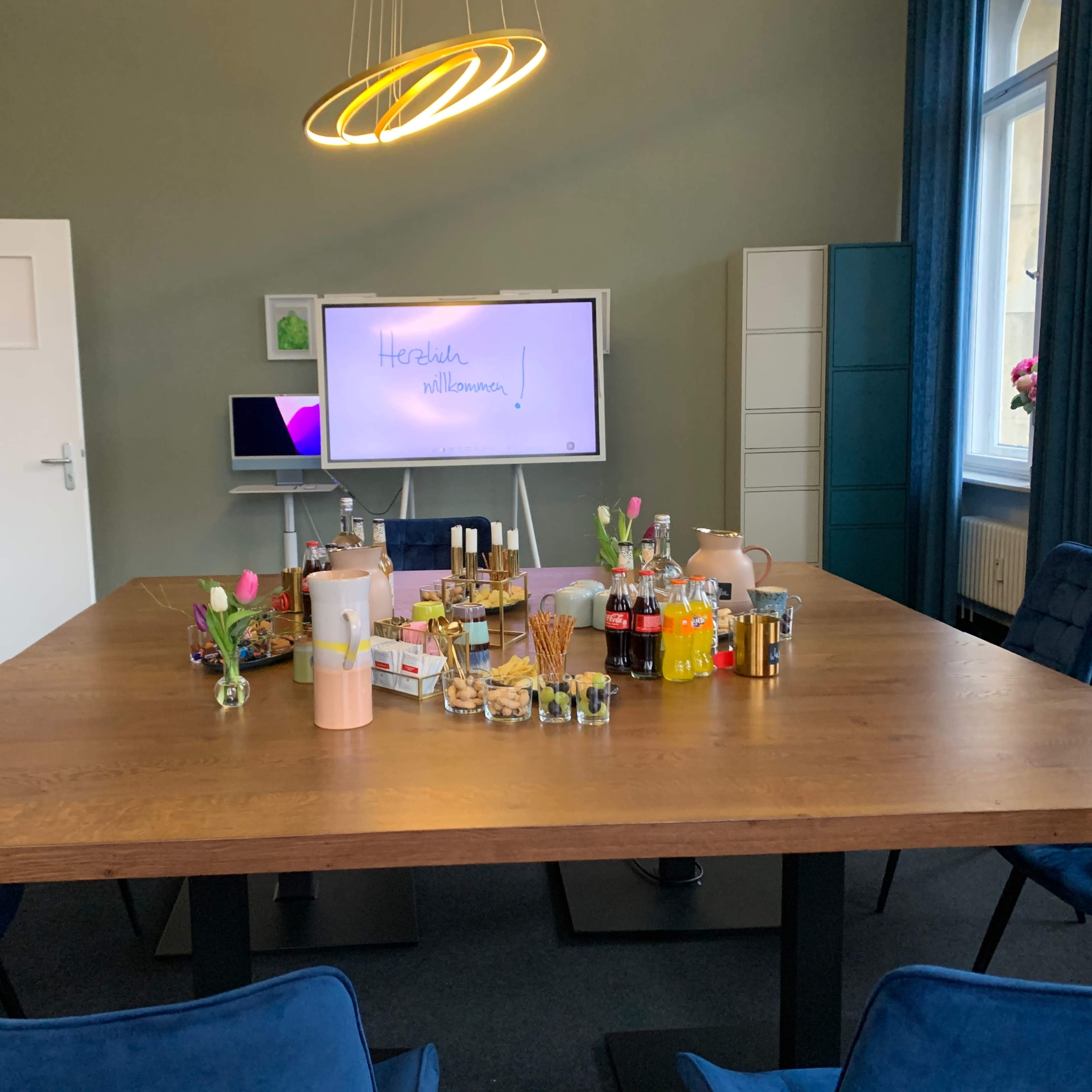 A conference room table is set up with various snacks and beverages, surrounded by blue chairs, and a presentation screen displays "Herzlich willkommen."