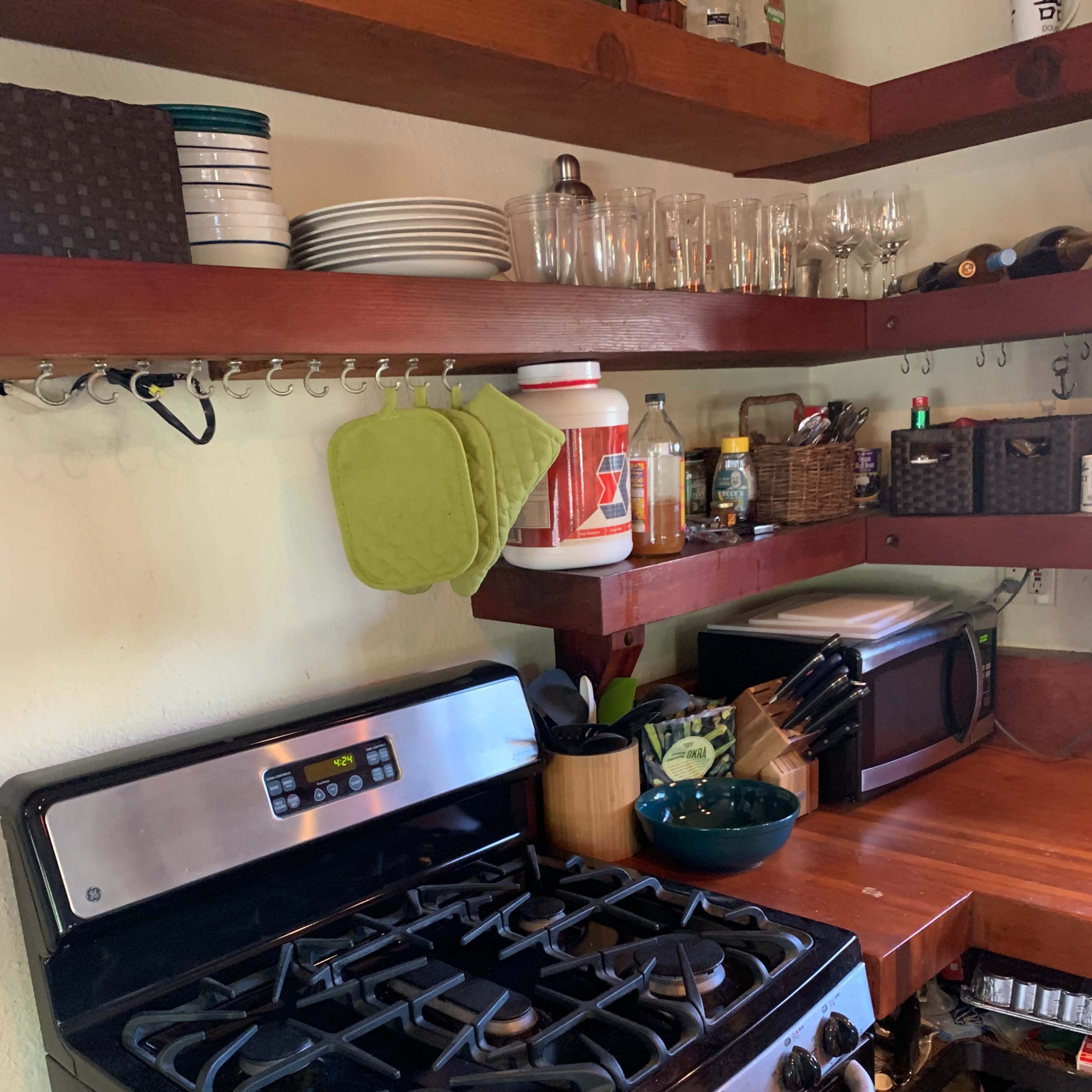 A kitchen with a gas stovetop, wooden shelves holding dishes and glasses, and a microwave on the countertop.