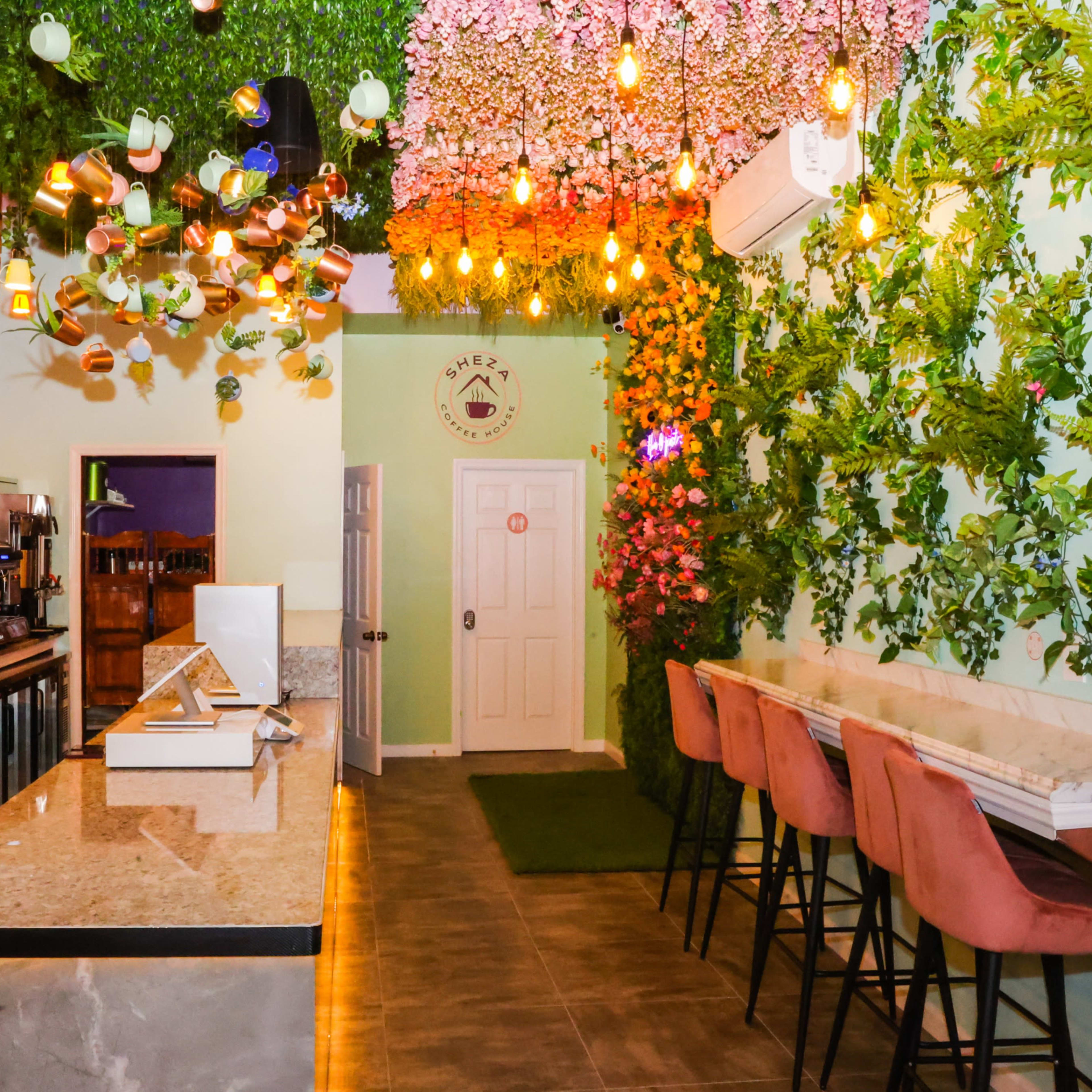 A modern café features a counter with a marble top, decorated walls covered in greenery and flowers, and colorful hanging light fixtures.