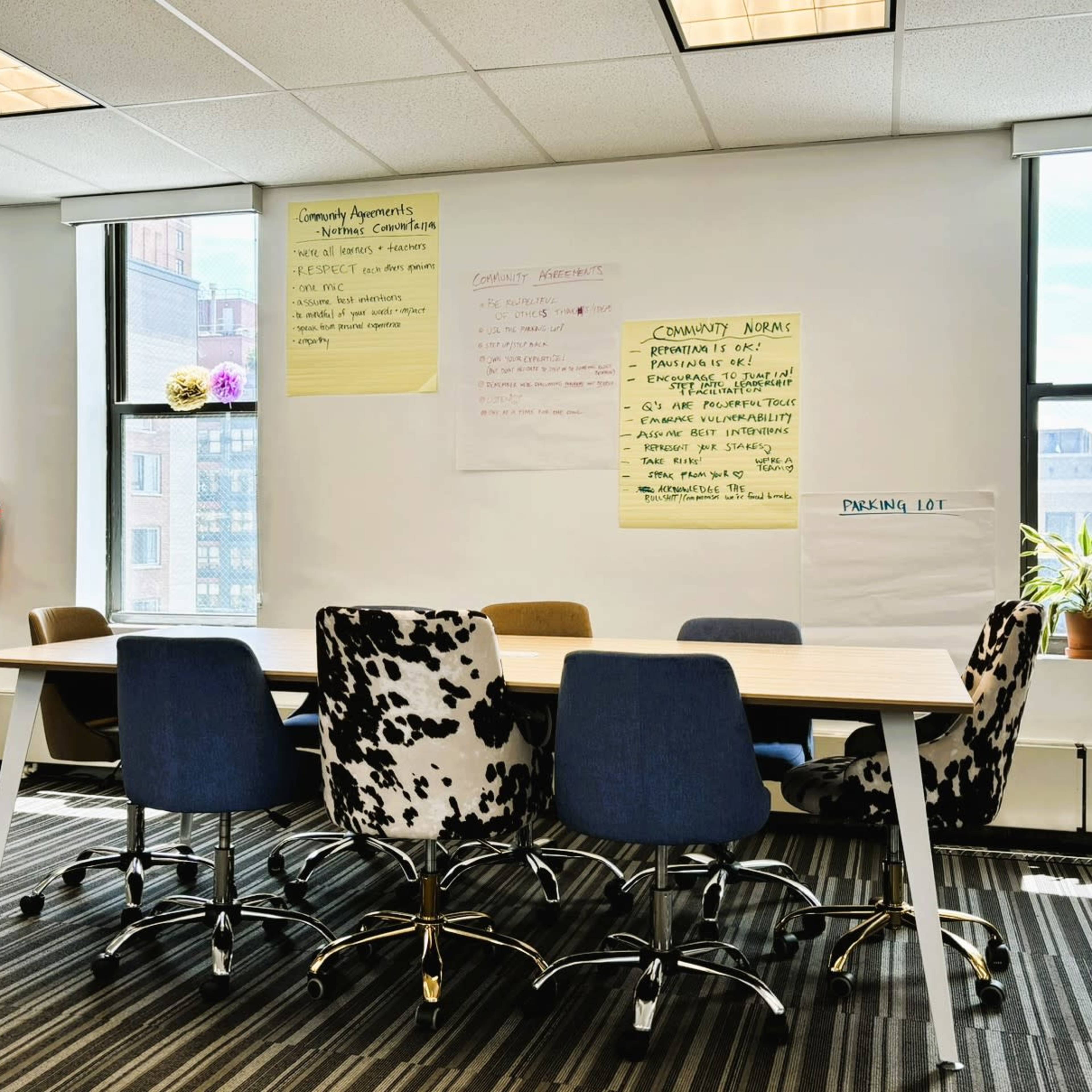 A meeting room features a long table surrounded by six chairs, with large windows and flip chart paper on the walls.
