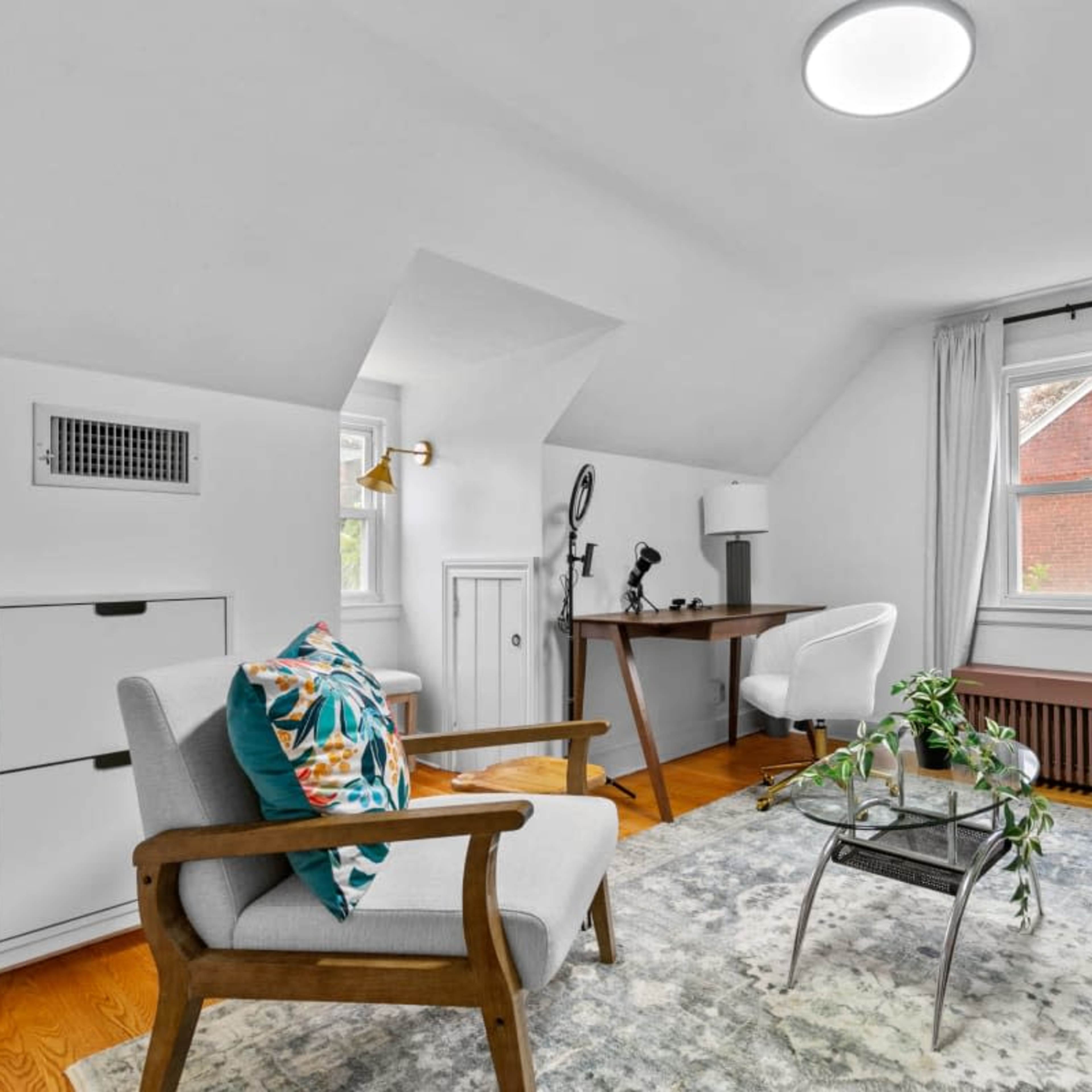 A bright, minimalist room featuring a seating area with a chair and sofa, a small desk, and a large window that provides natural light.