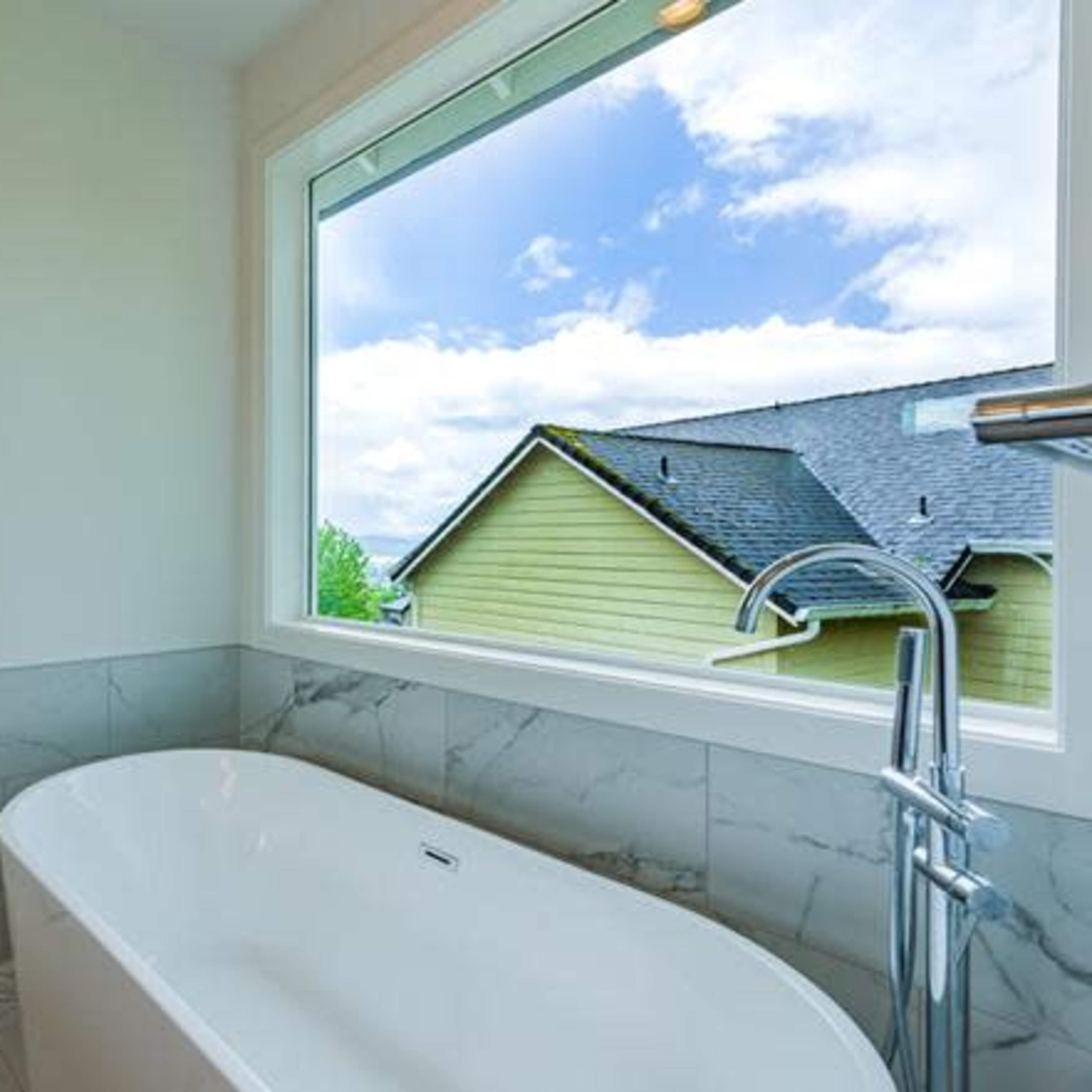 A modern bathroom features a freestanding bathtub next to a large window with a view of a green landscape and houses.
