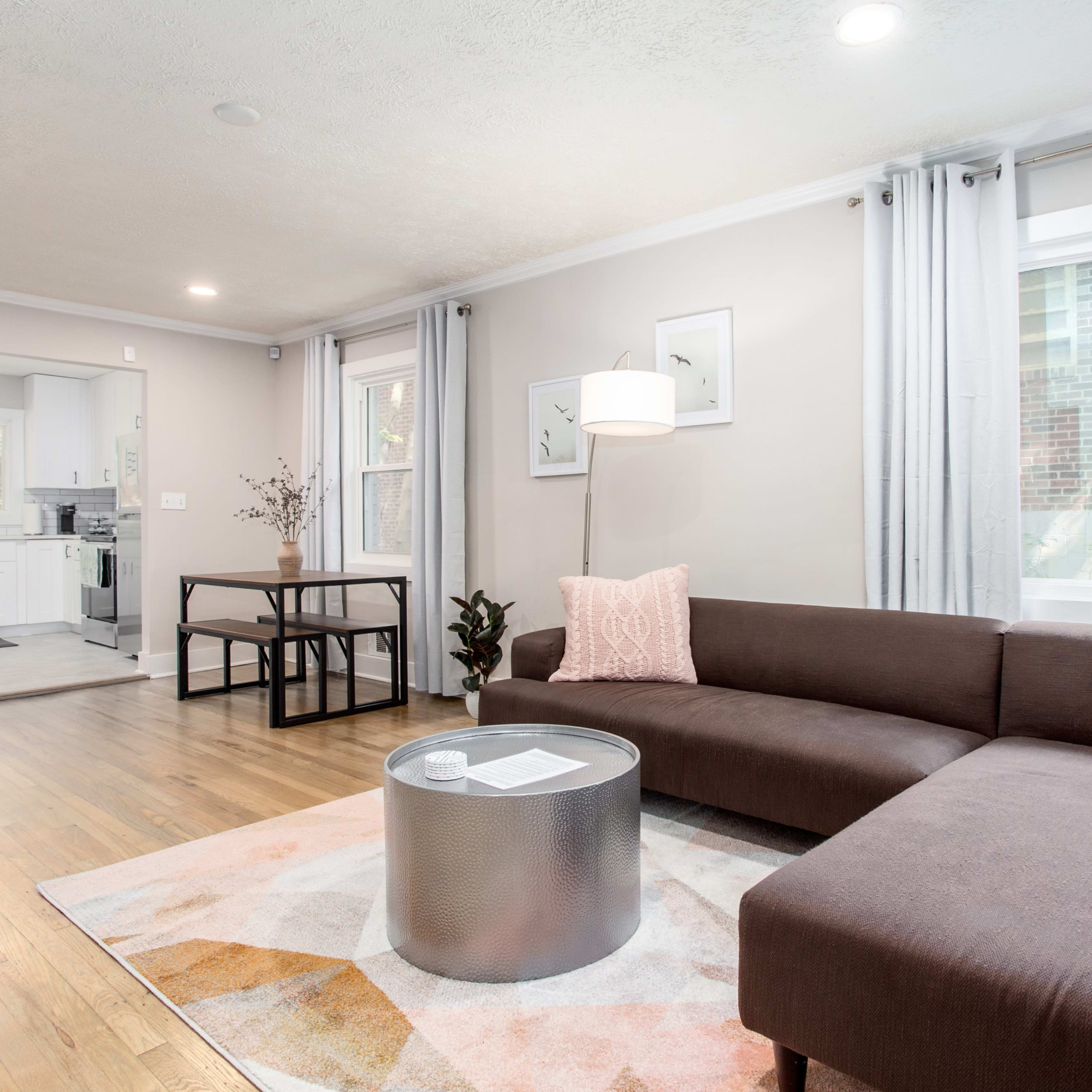 A modern living room features a brown sectional sofa, a round silver coffee table, and a small dining area, with large windows allowing natural light to fill the space.