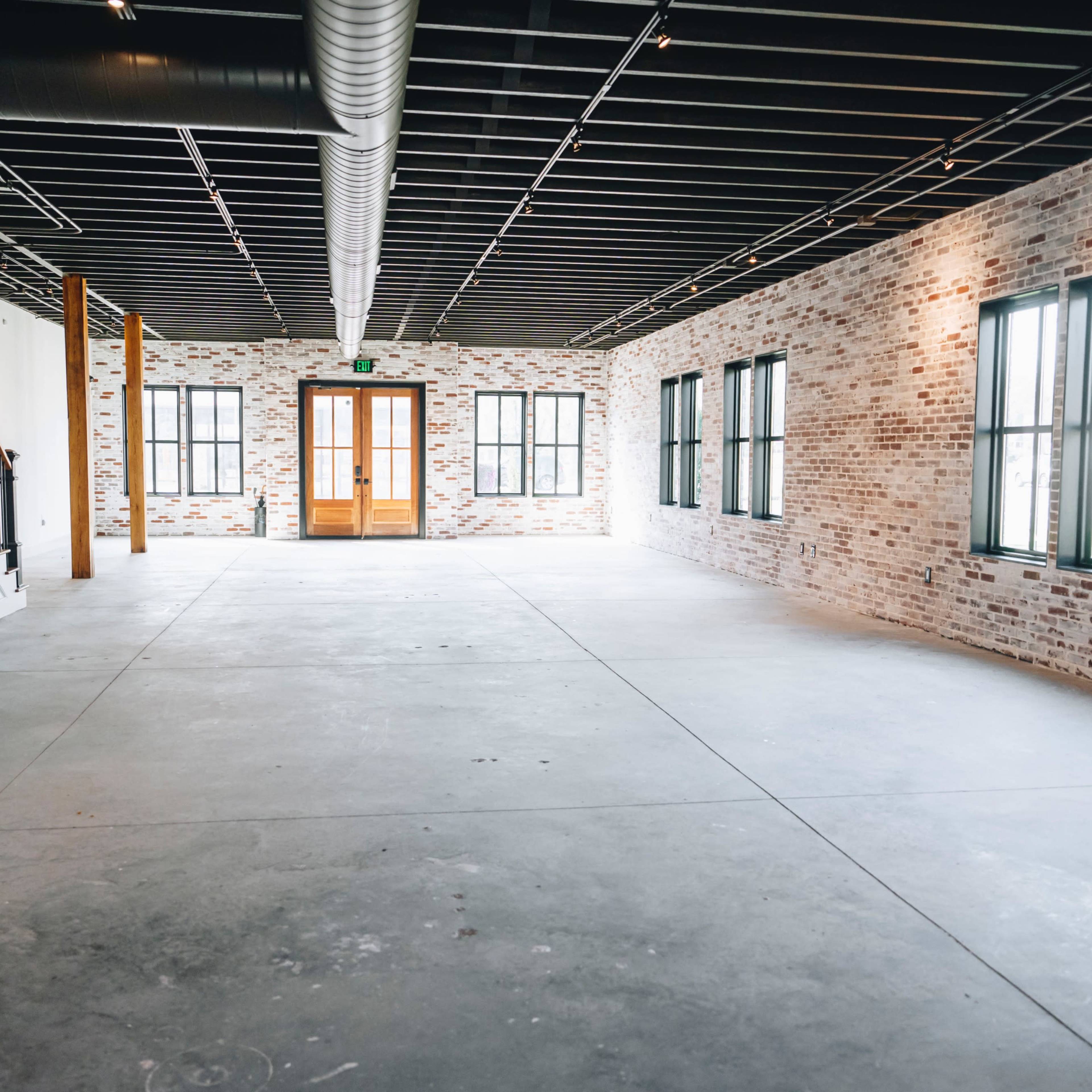 An empty interior space with exposed brick walls, large windows, and a concrete floor.