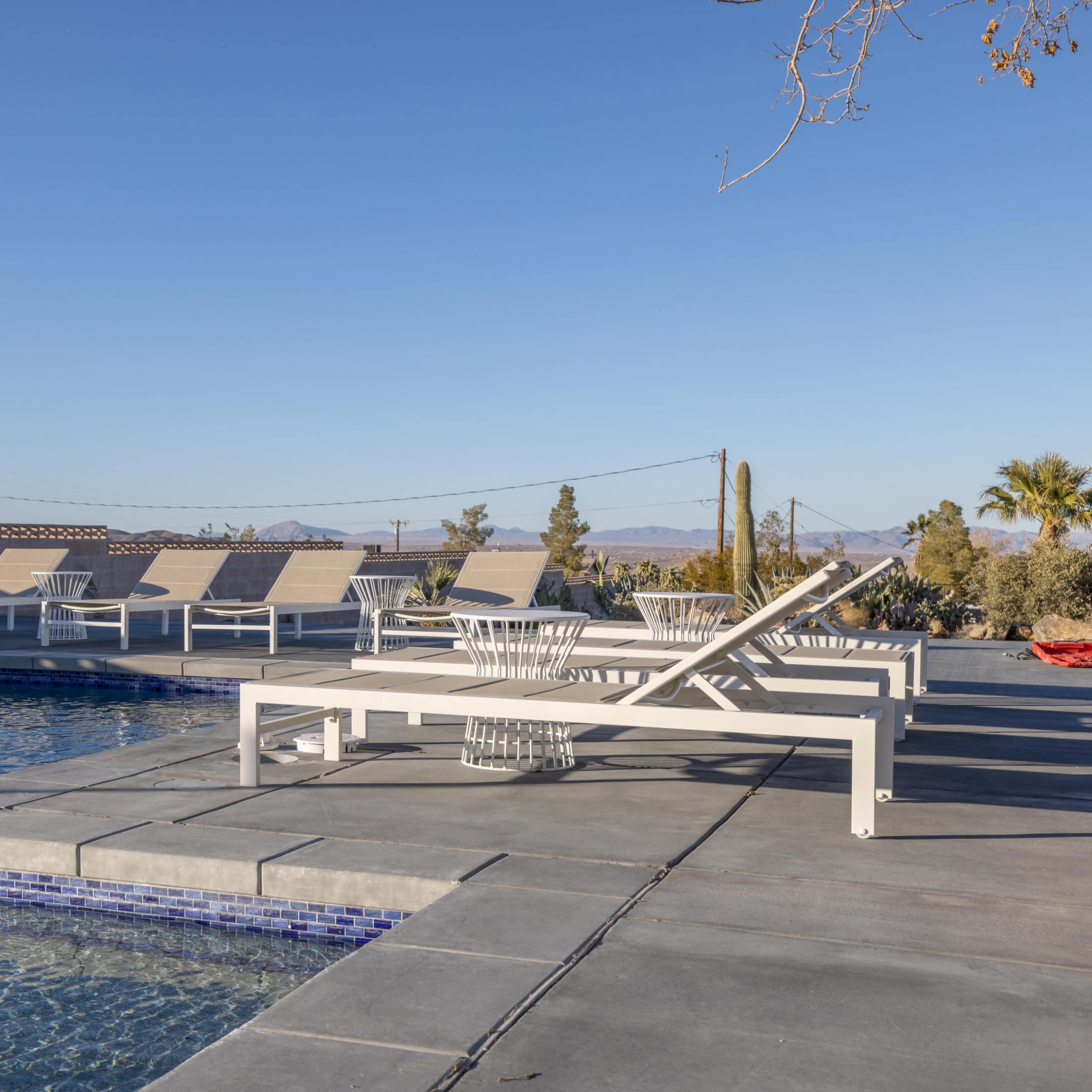 A modern swimming pool area with several white lounge chairs arranged around it, surrounded by sparse desert landscaping.
