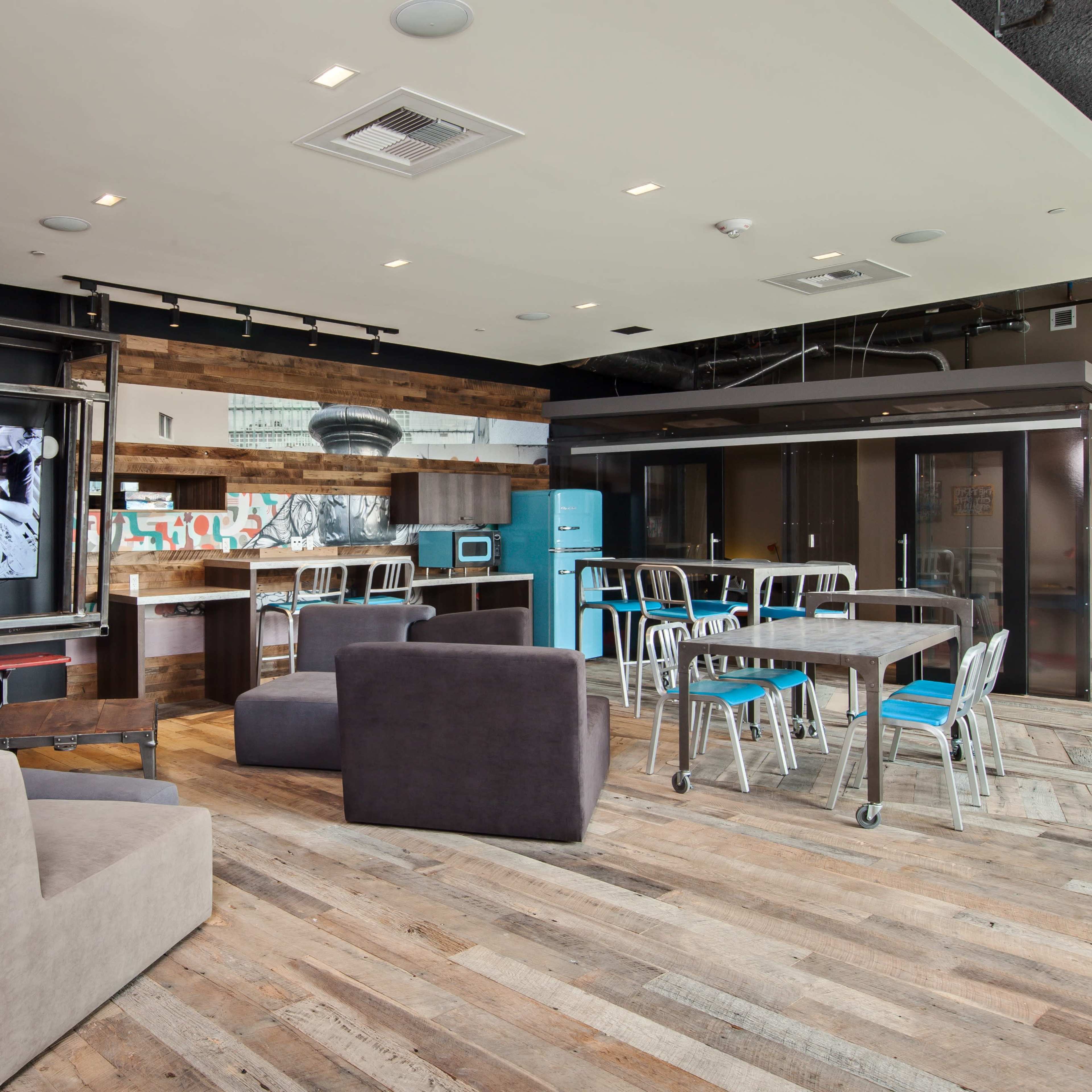 A modern communal space featuring a kitchen area, a large television, and various seating options arranged around a wooden floor.