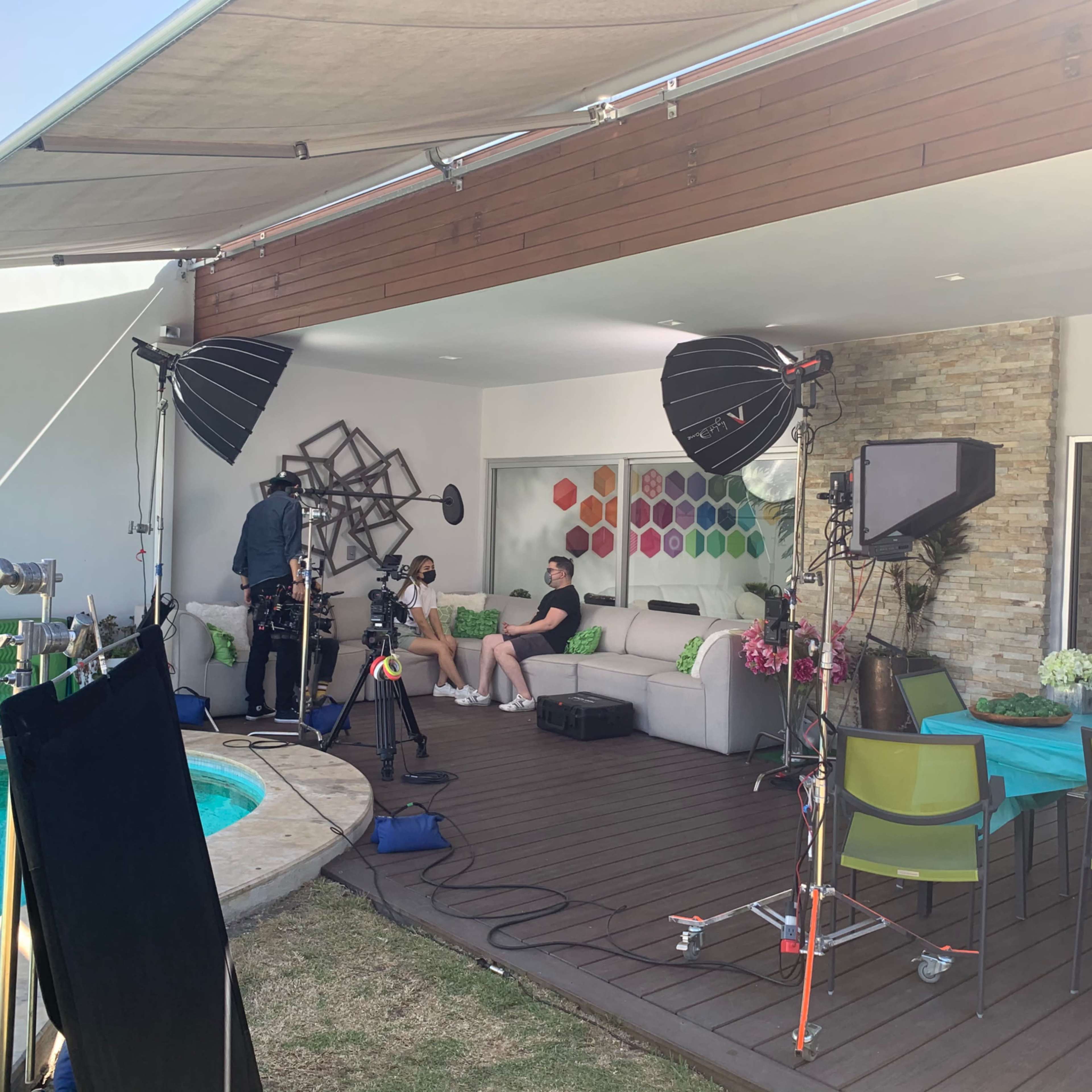 A film set outdoors near a swimming pool, featuring lighting equipment, cameras, and crew members preparing for a shoot with two people sitting on a couch.
