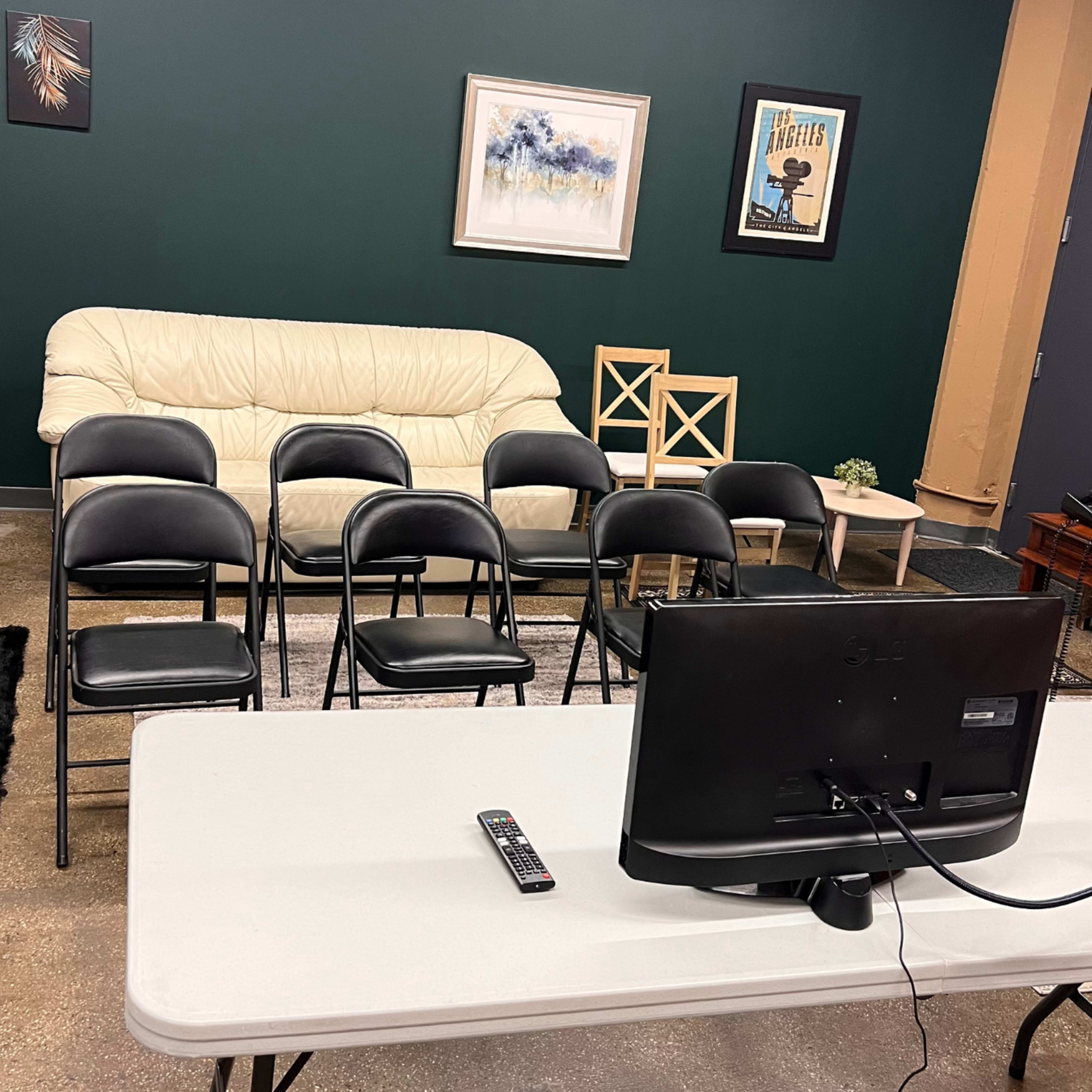 A room with a light-colored couch, several black chairs arranged in rows, a television on a table, and a framed picture on the wall.