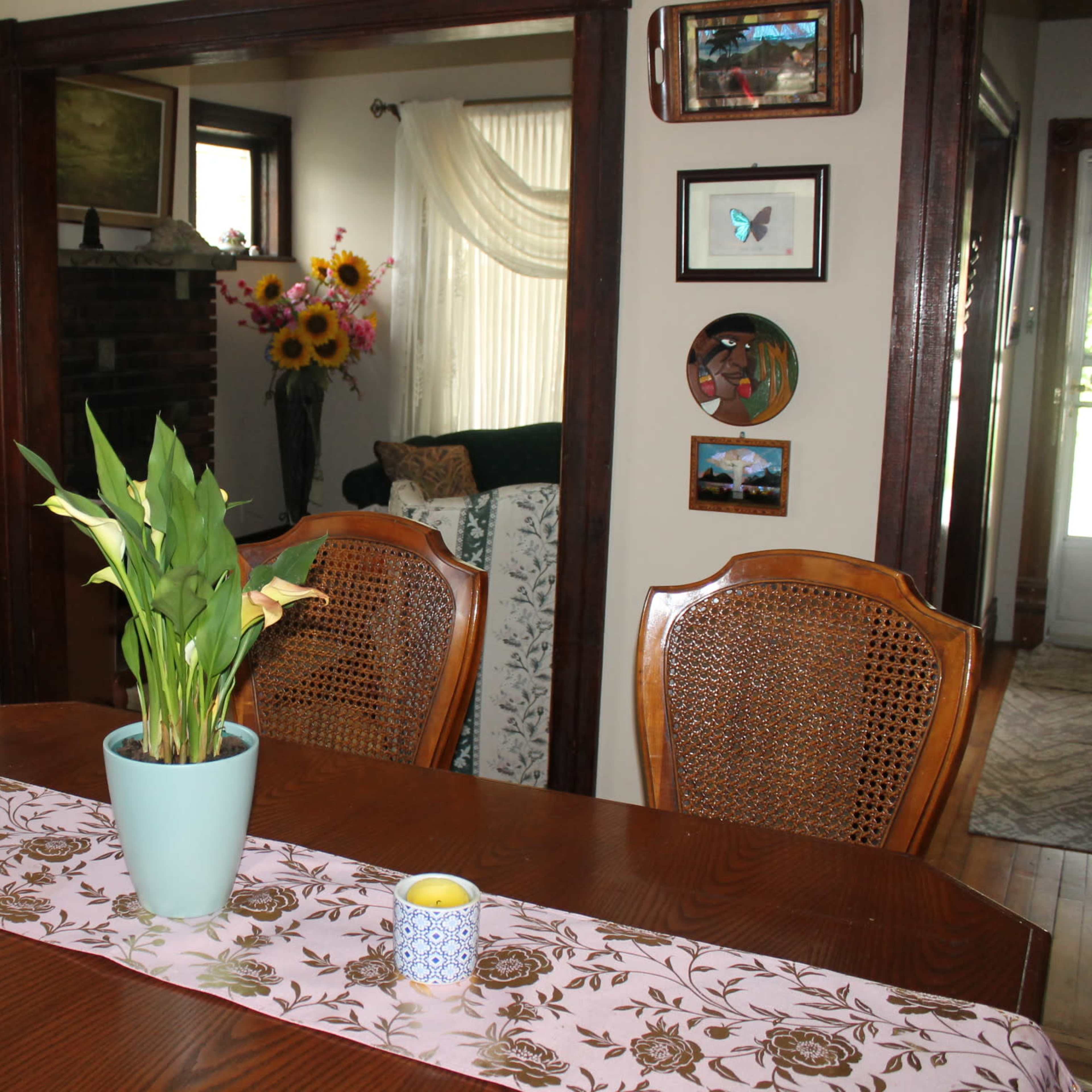 A wooden dining table with a pink table runner and a potted plant is set in a room adorned with art on the walls and a view into a living area.