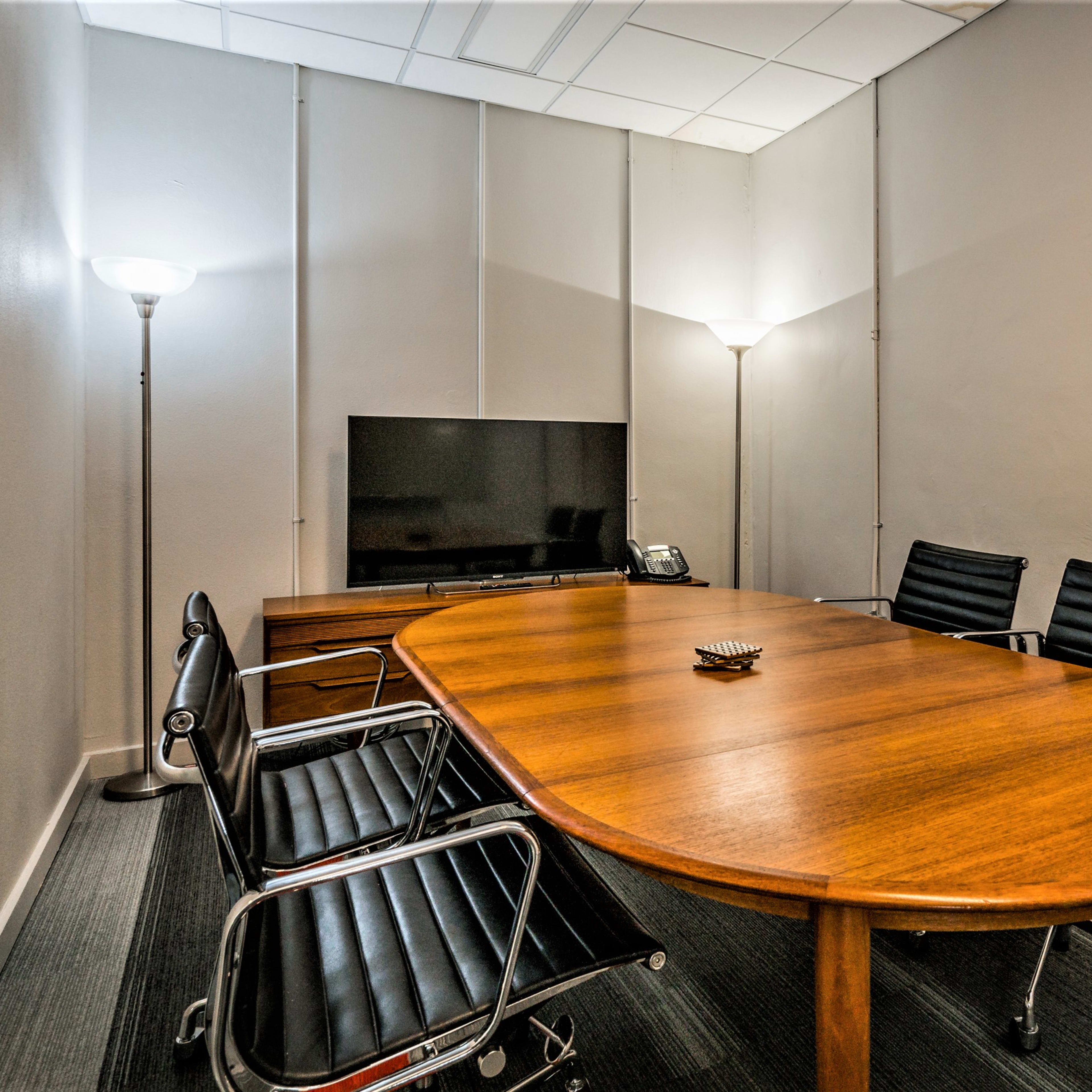 A small conference room features a wooden table surrounded by four black chairs, a television on the wall, and two floor lamps.