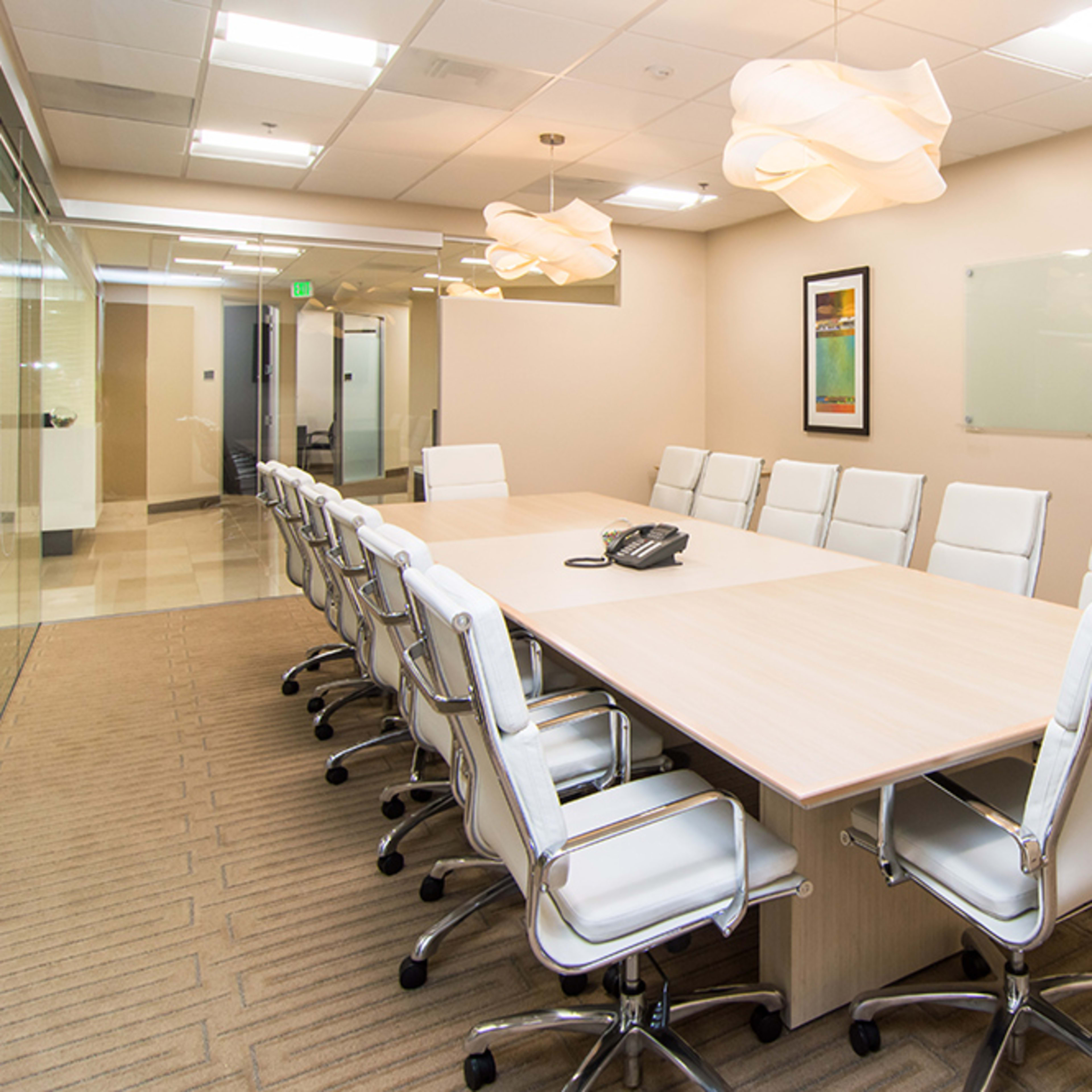 A long conference table with white chairs is set up in a well-lit meeting room featuring modern lighting and artwork on the walls.
