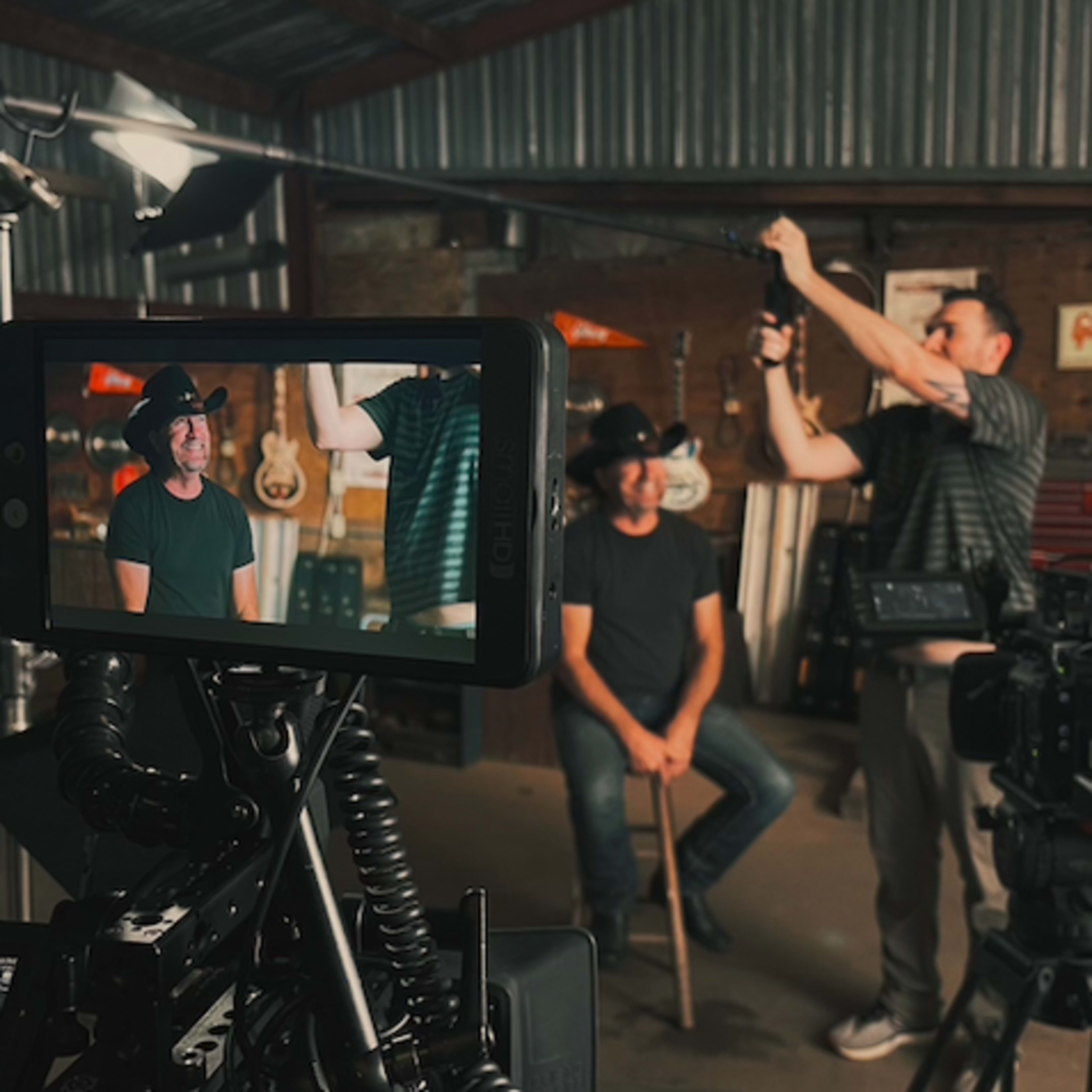 A camera crew films an interview with a seated man holding a cane in a rustic studio filled with guitars.