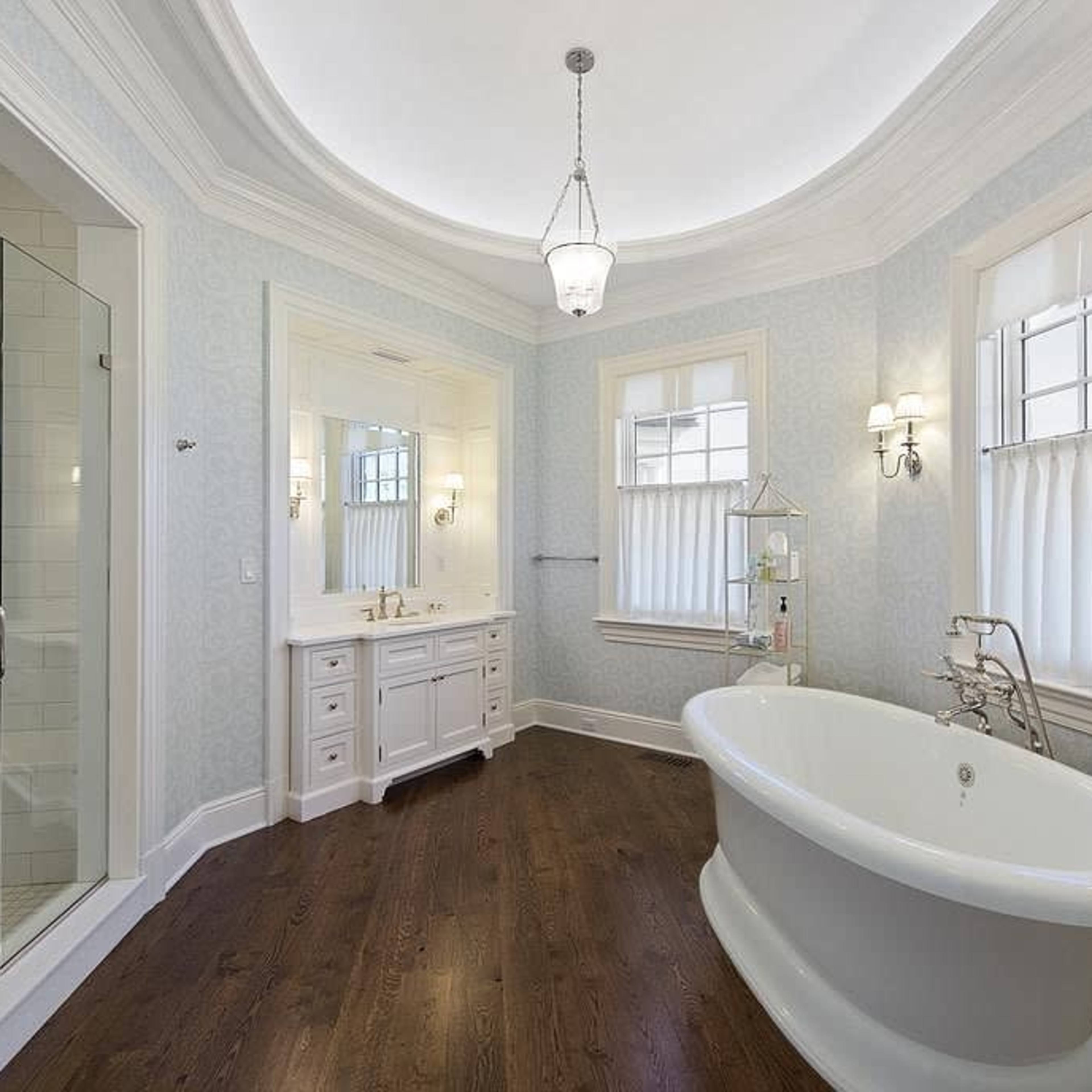 A spacious bathroom featuring a freestanding bathtub, a glass shower, and dual sinks with a wooden floor and light-colored walls.