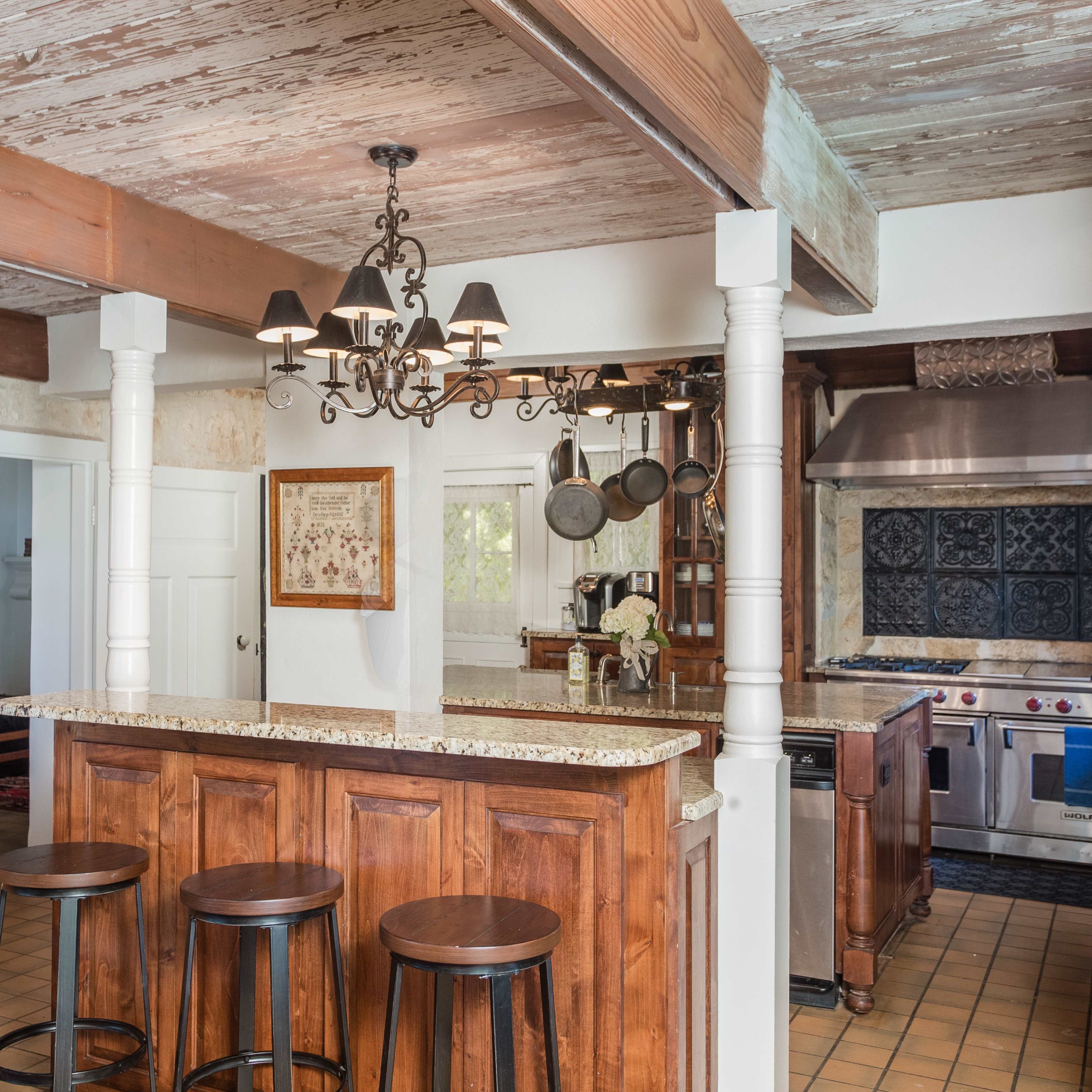 A kitchen with a granite countertop island, bar stools, wooden cabinets, and stainless steel appliances under a chandelier.