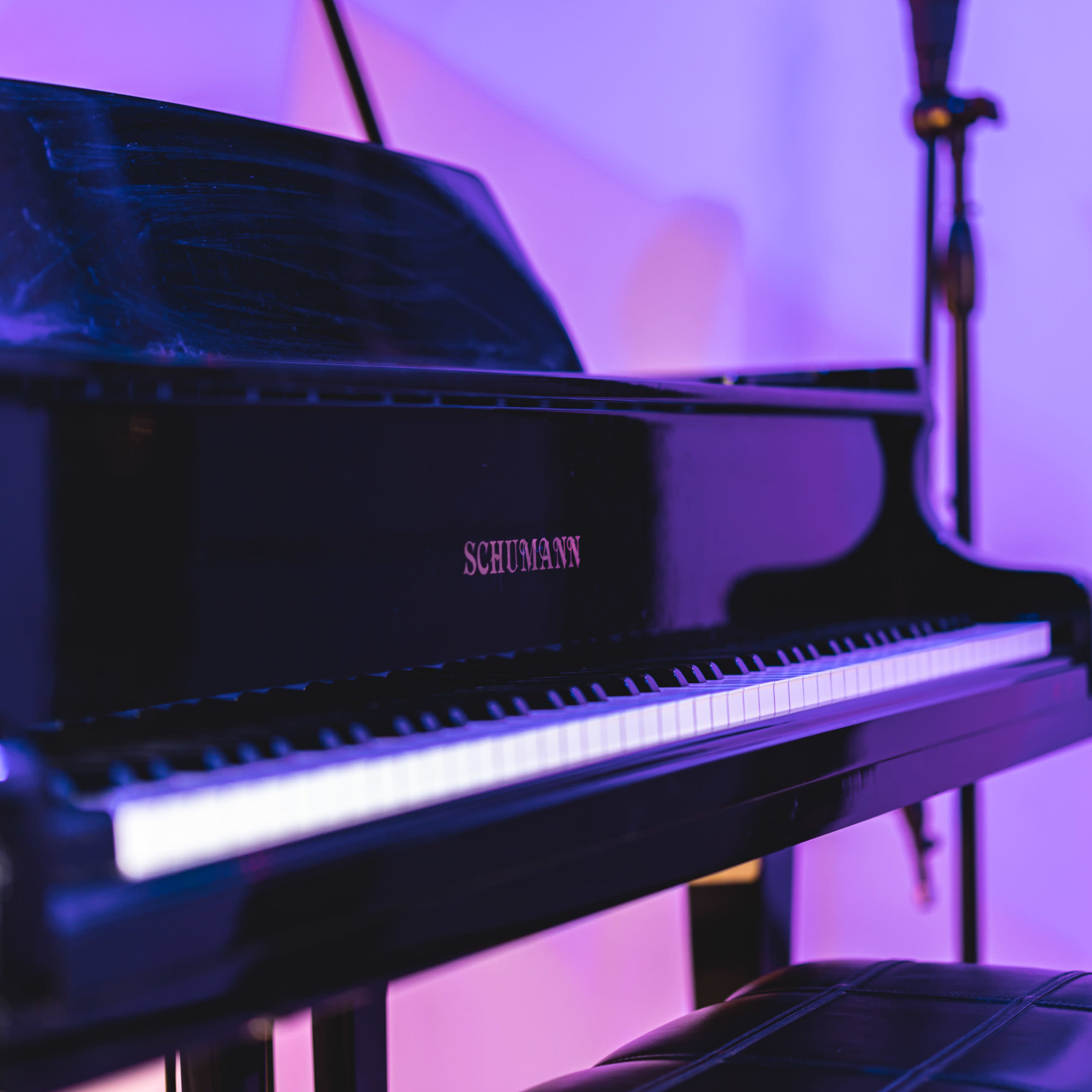 A black grand piano is positioned beside a microphone stand under colored lighting.