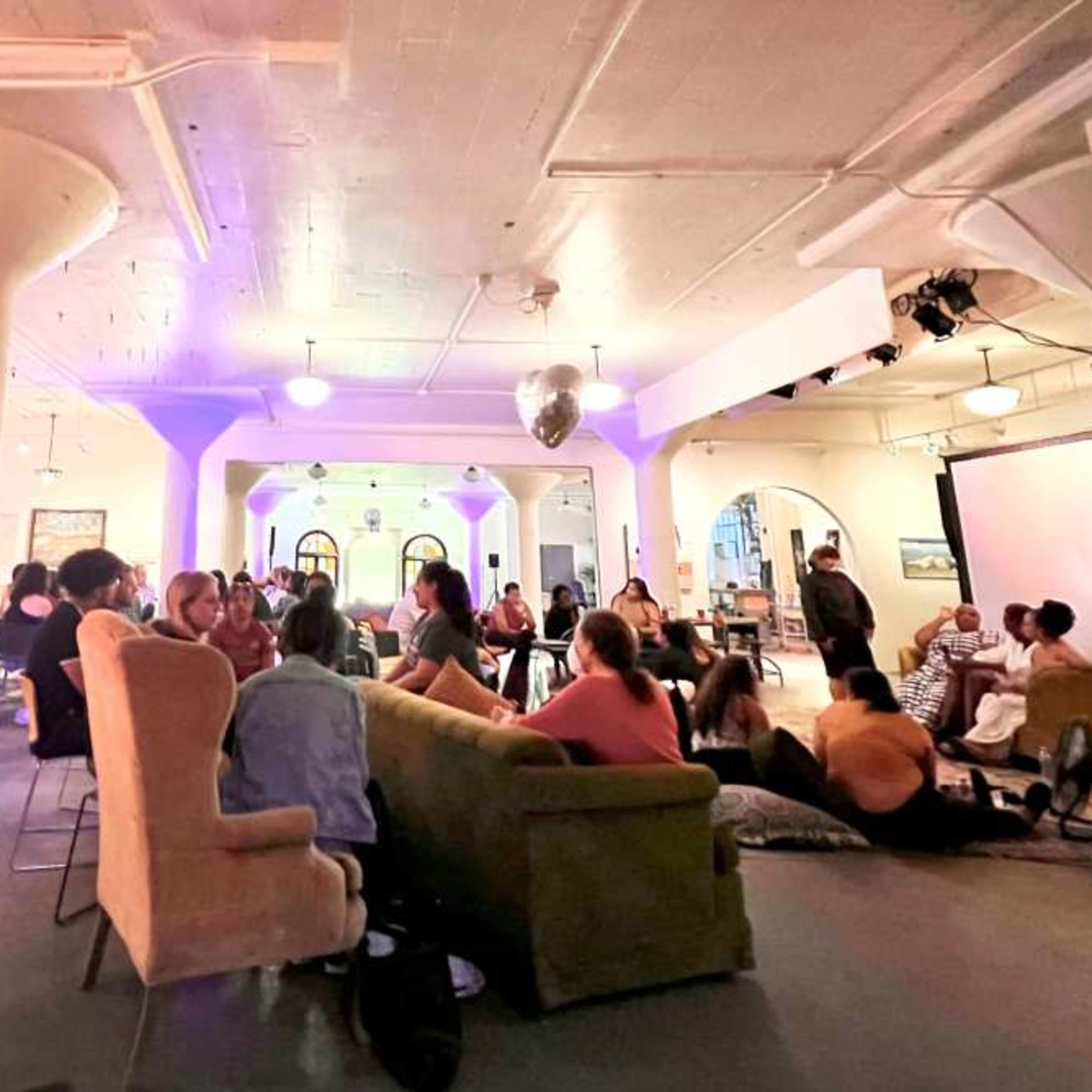 A group of people gathers in a spacious room with purple lighting, chairs arranged in various configurations, and decorative pillars.