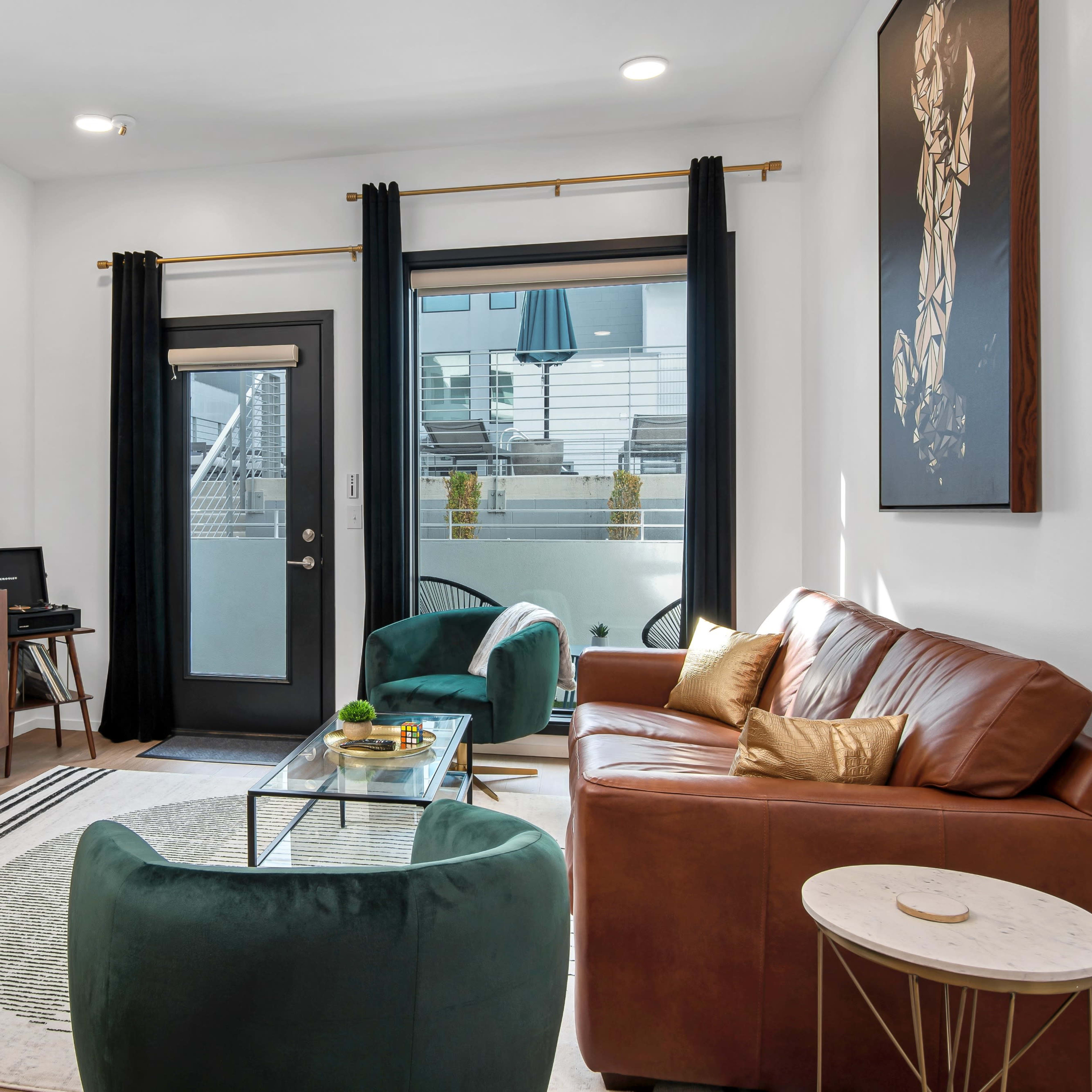The living room features a brown leather sofa, green accent chairs, a glass coffee table, and a large wall-mounted TV, with floor-to-ceiling windows and black curtains.