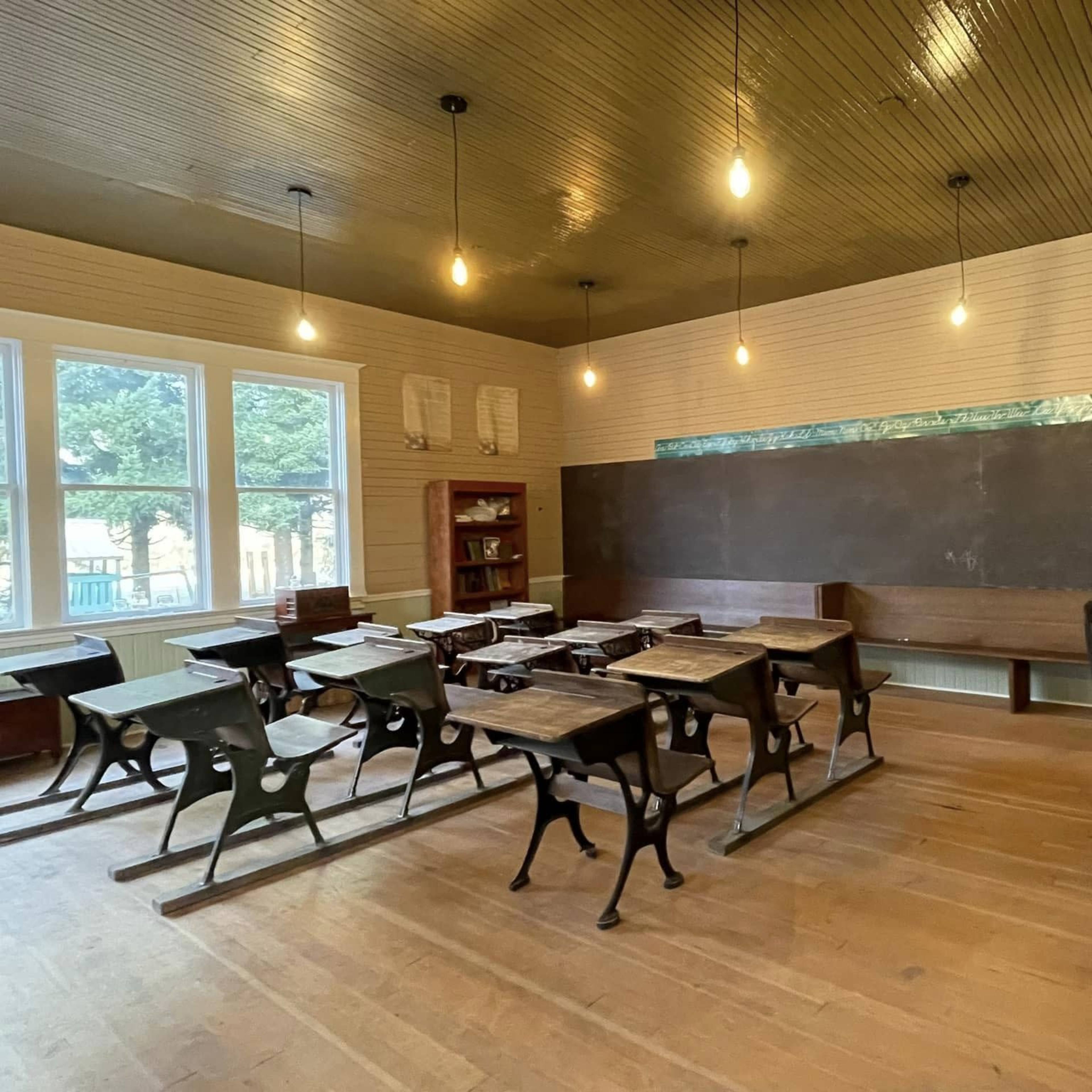 A vintage classroom contains wooden desks, a chalkboard, and a bookshelf, illuminated by hanging light bulbs.