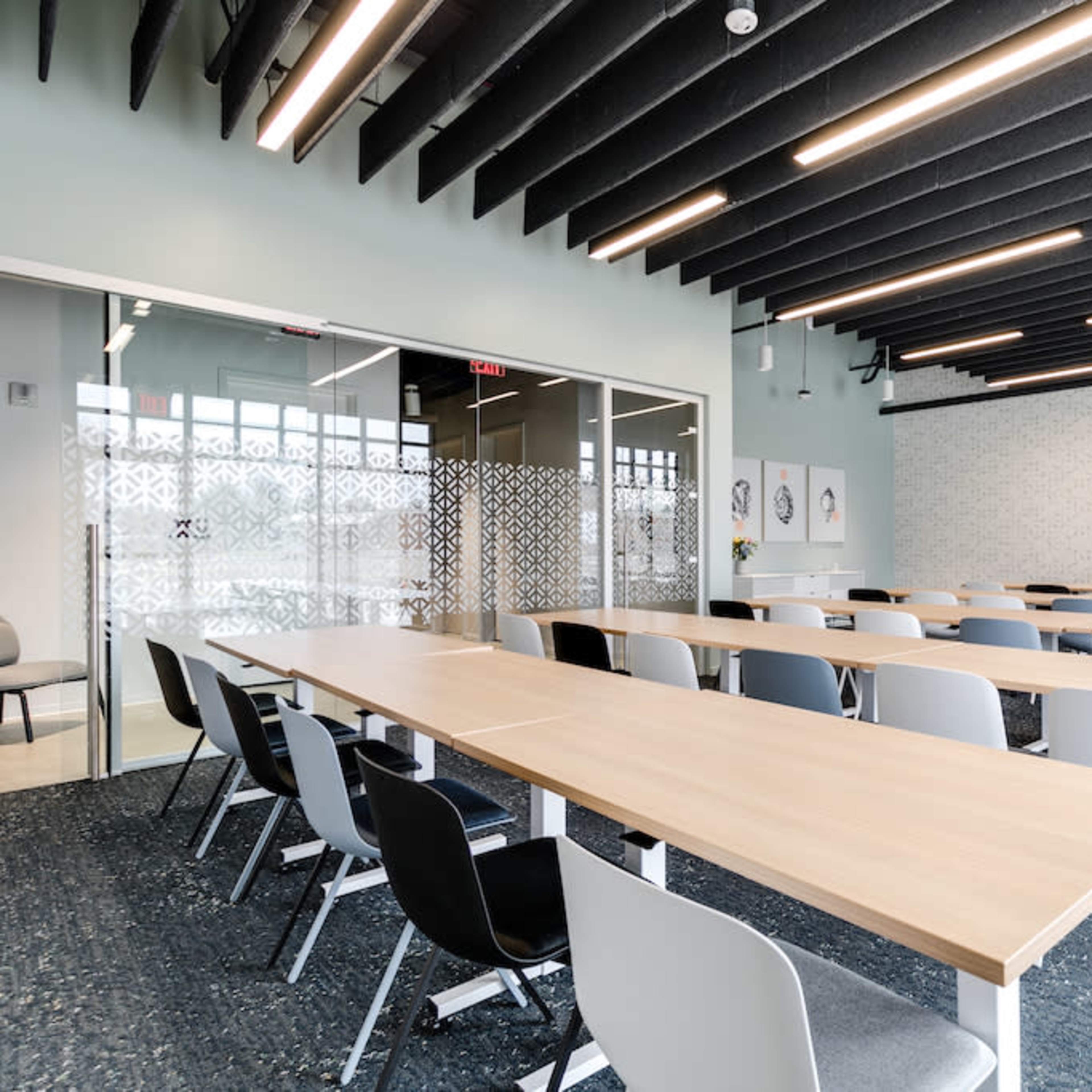 A modern conference room features long tables arranged in rows with chairs, large windows, and a wall-mounted screen.