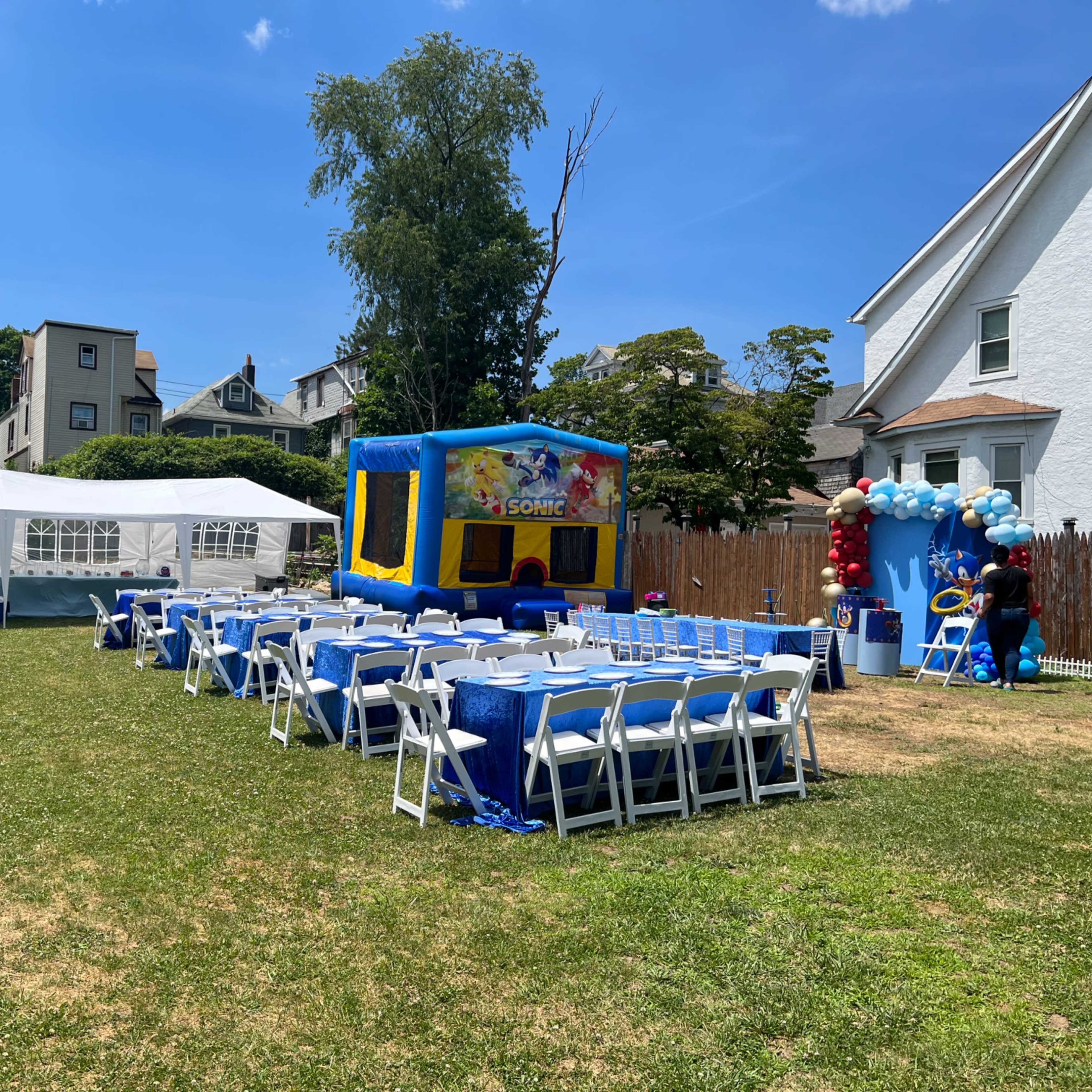 A backyard is set up for a celebration with a large inflatable bounce house, tables arranged with blue table covers, and a tent in the background.
