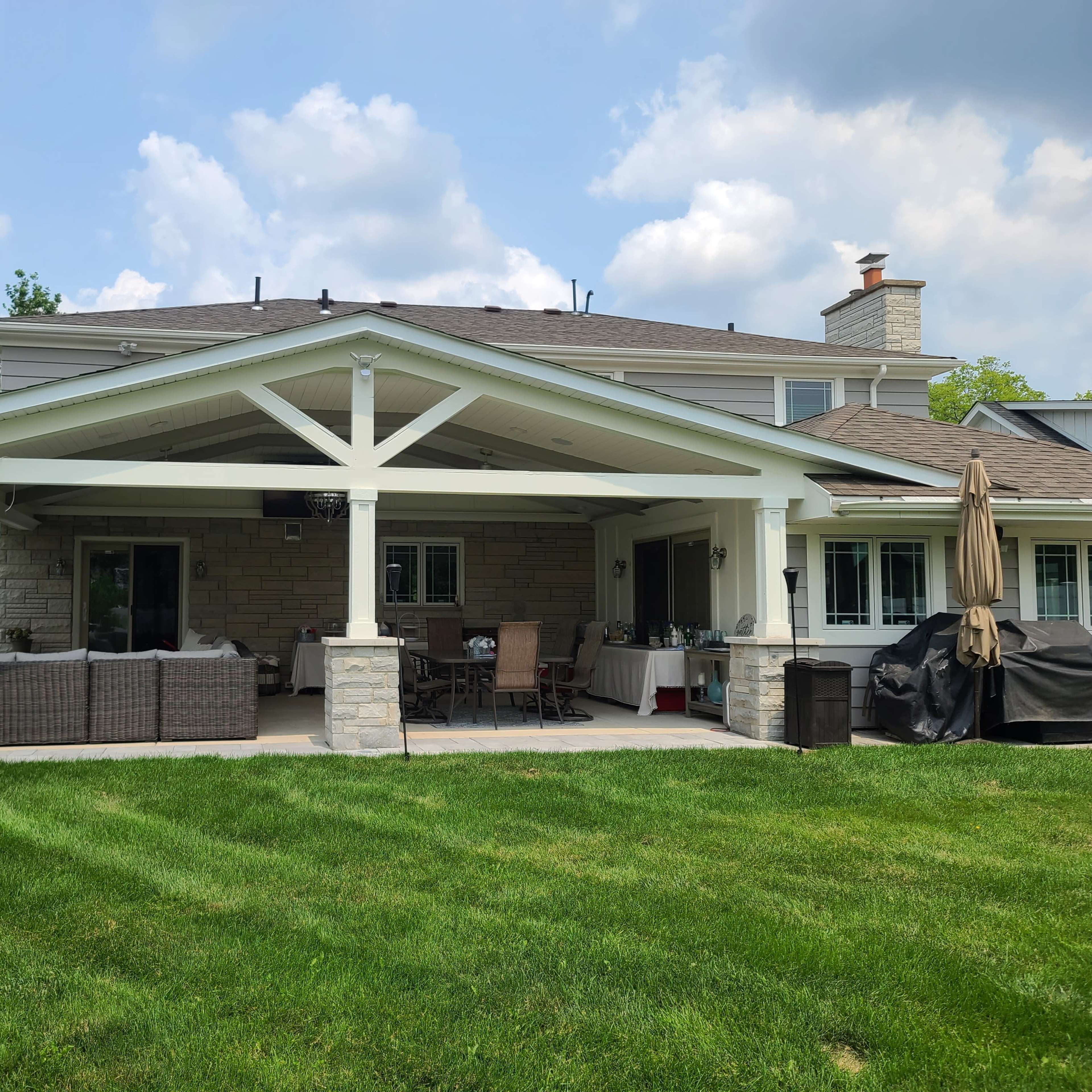 A backyard patio with a large covered area featuring stone pillars, outdoor furniture, and a well-maintained lawn under a partly cloudy sky.