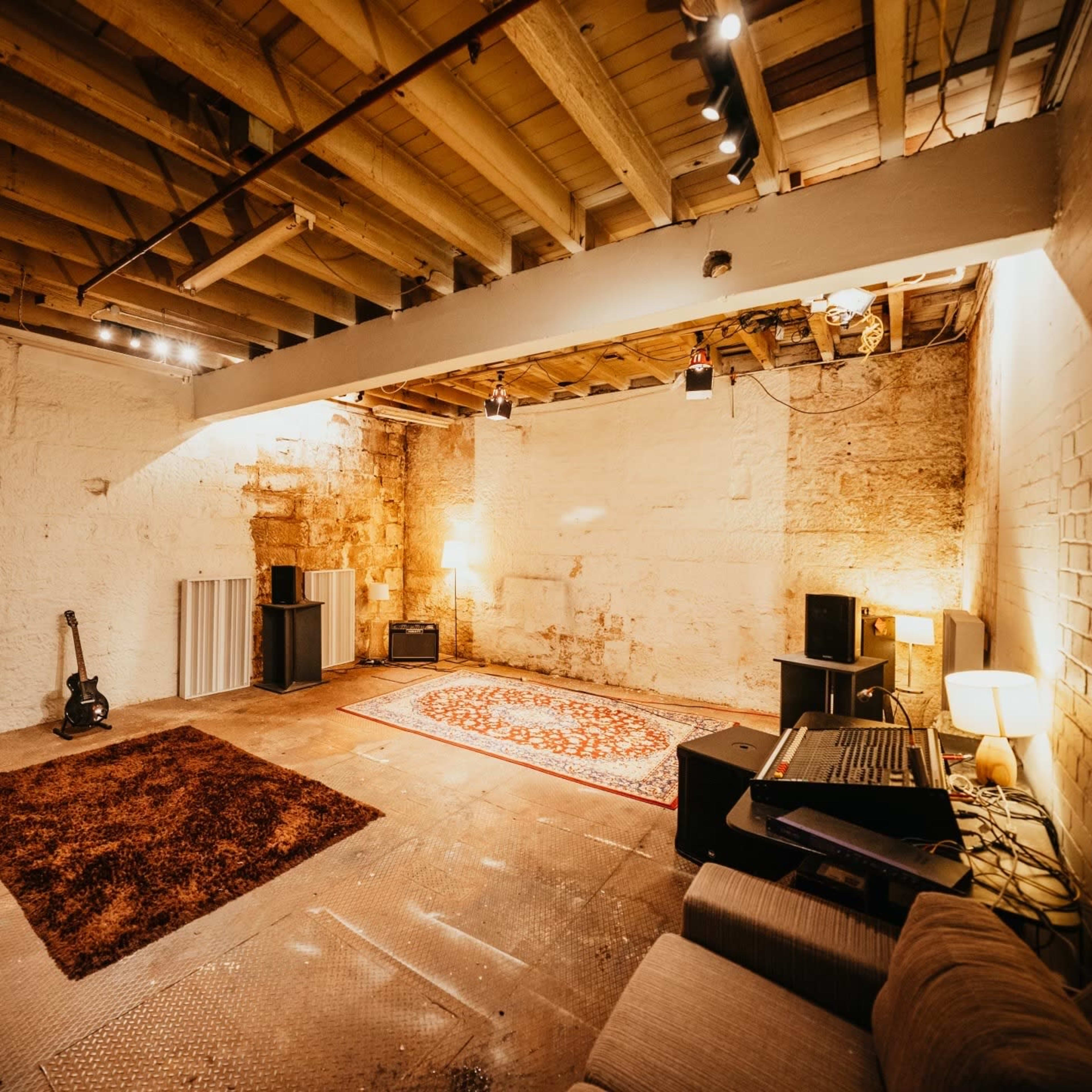 A spacious basement room features wooden beams, a patterned rug, various furniture pieces, and audio equipment along the walls.