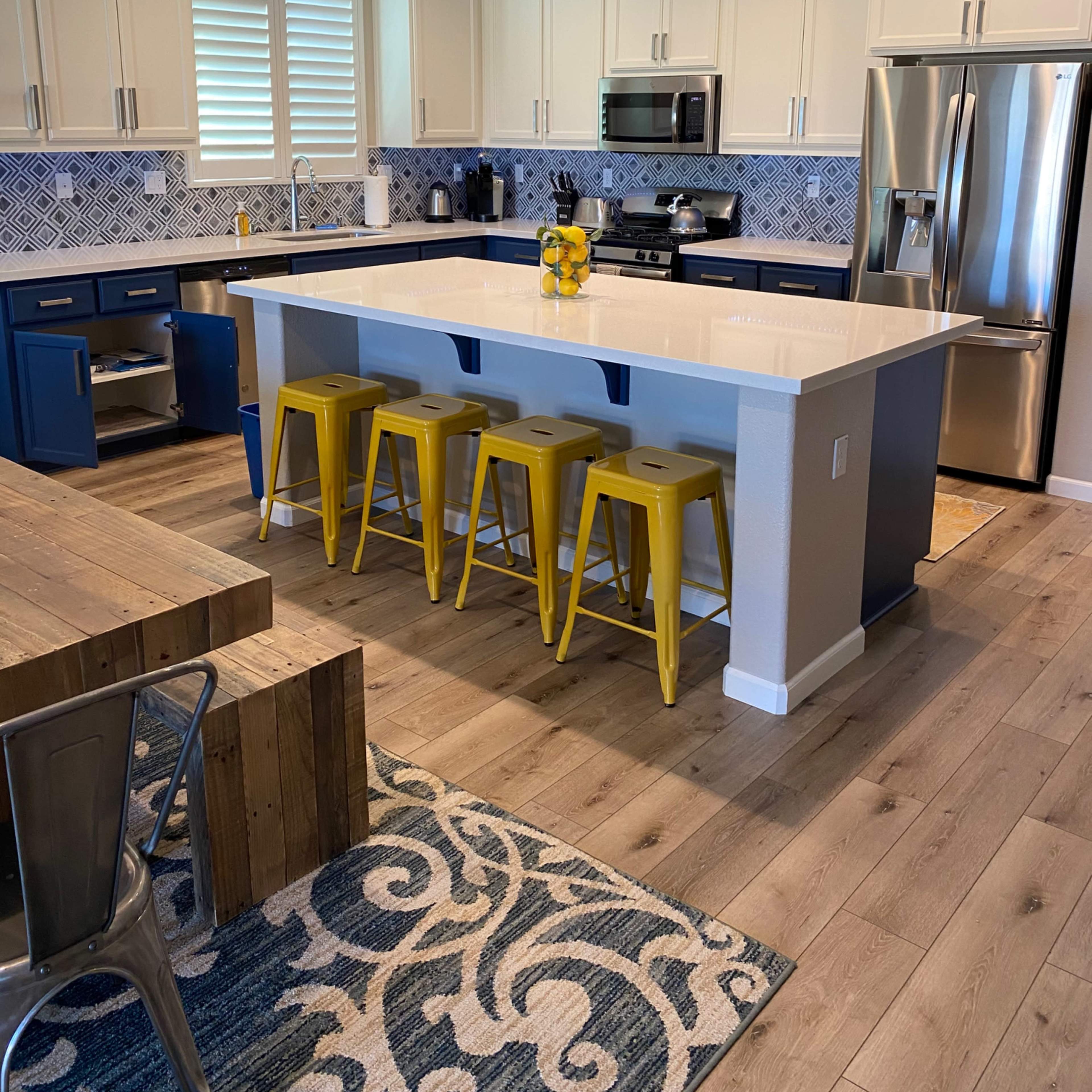 A modern kitchen features white cabinets, a blue island with yellow stools, stainless steel appliances, and a patterned rug.