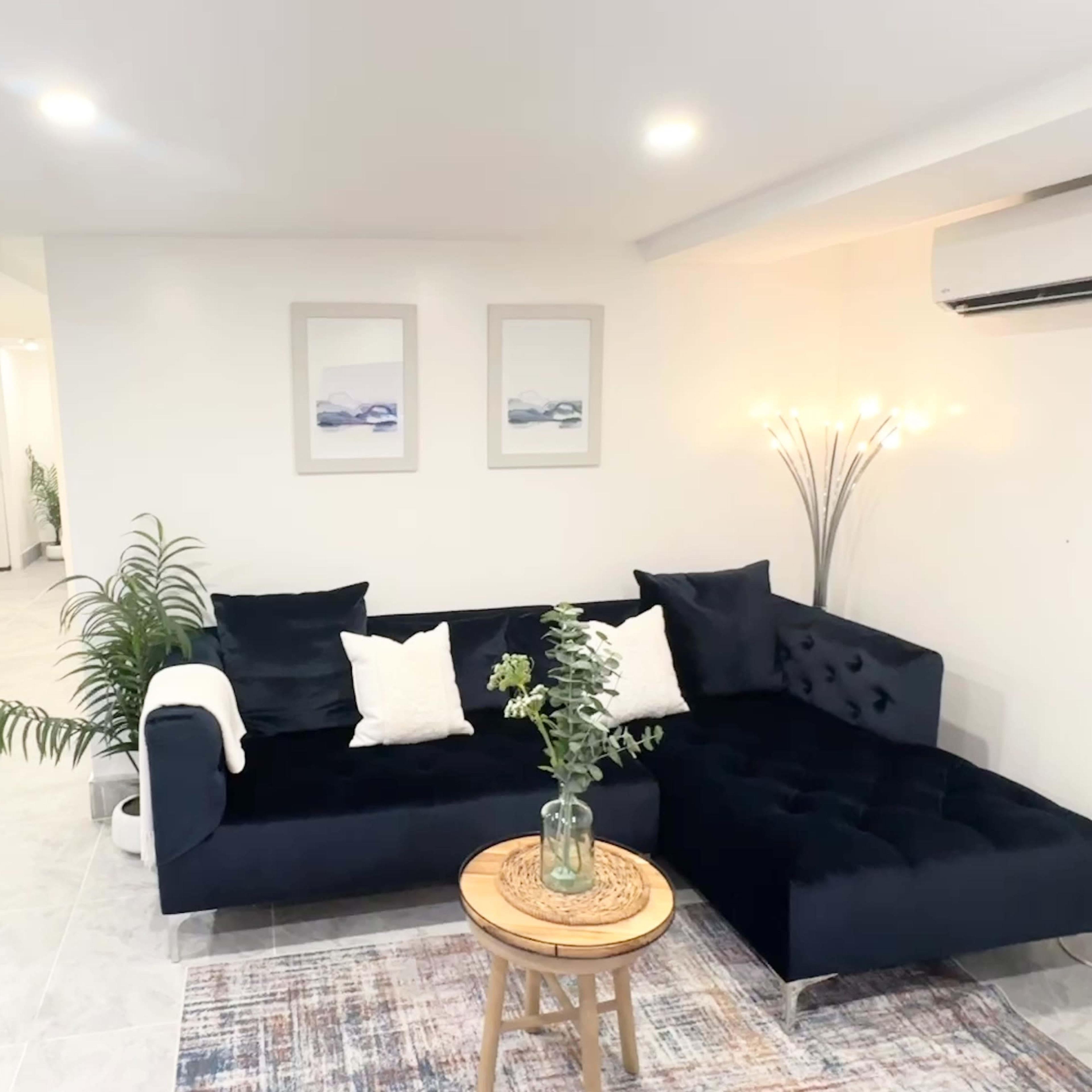 A modern living room features a dark sofa with decorative cushions, a round wooden table, and a plant, illuminated by soft overhead lighting.