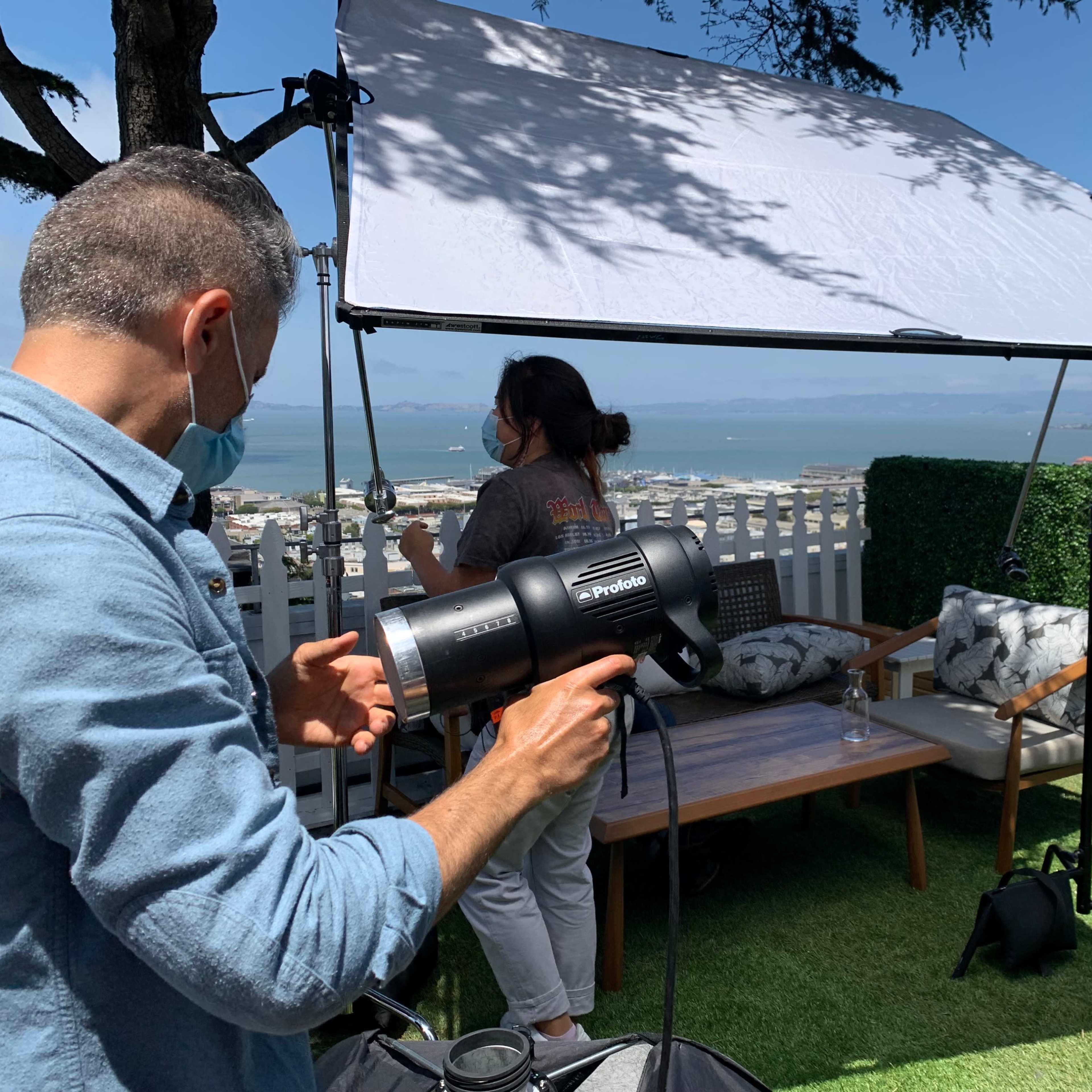 A crew sets up lighting equipment under a large diffuser near an outdoor seating area with a view of the landscape.