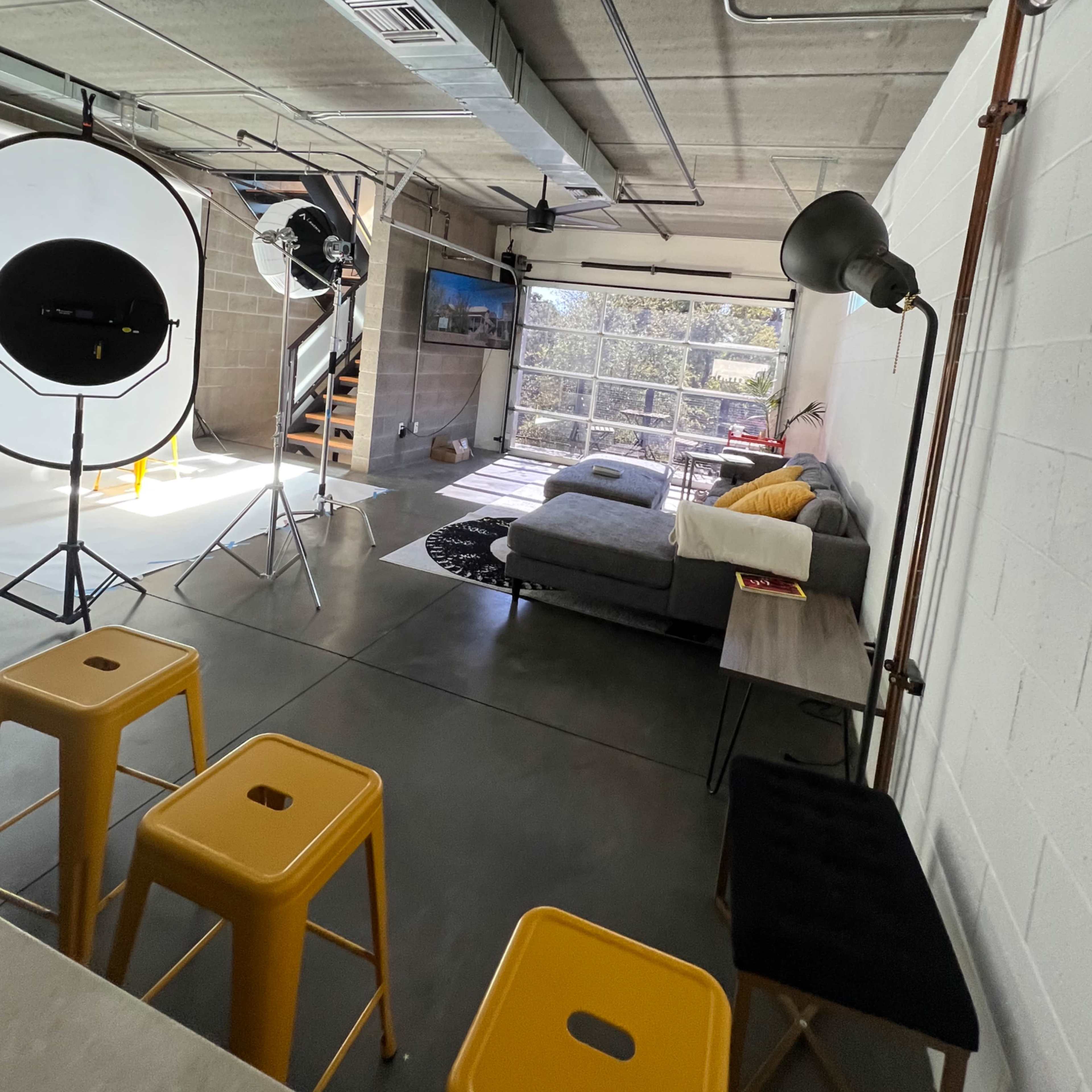 A modern, open-concept living space with a gray couch, lighting equipment, and a set of bar stools in a well-lit area.