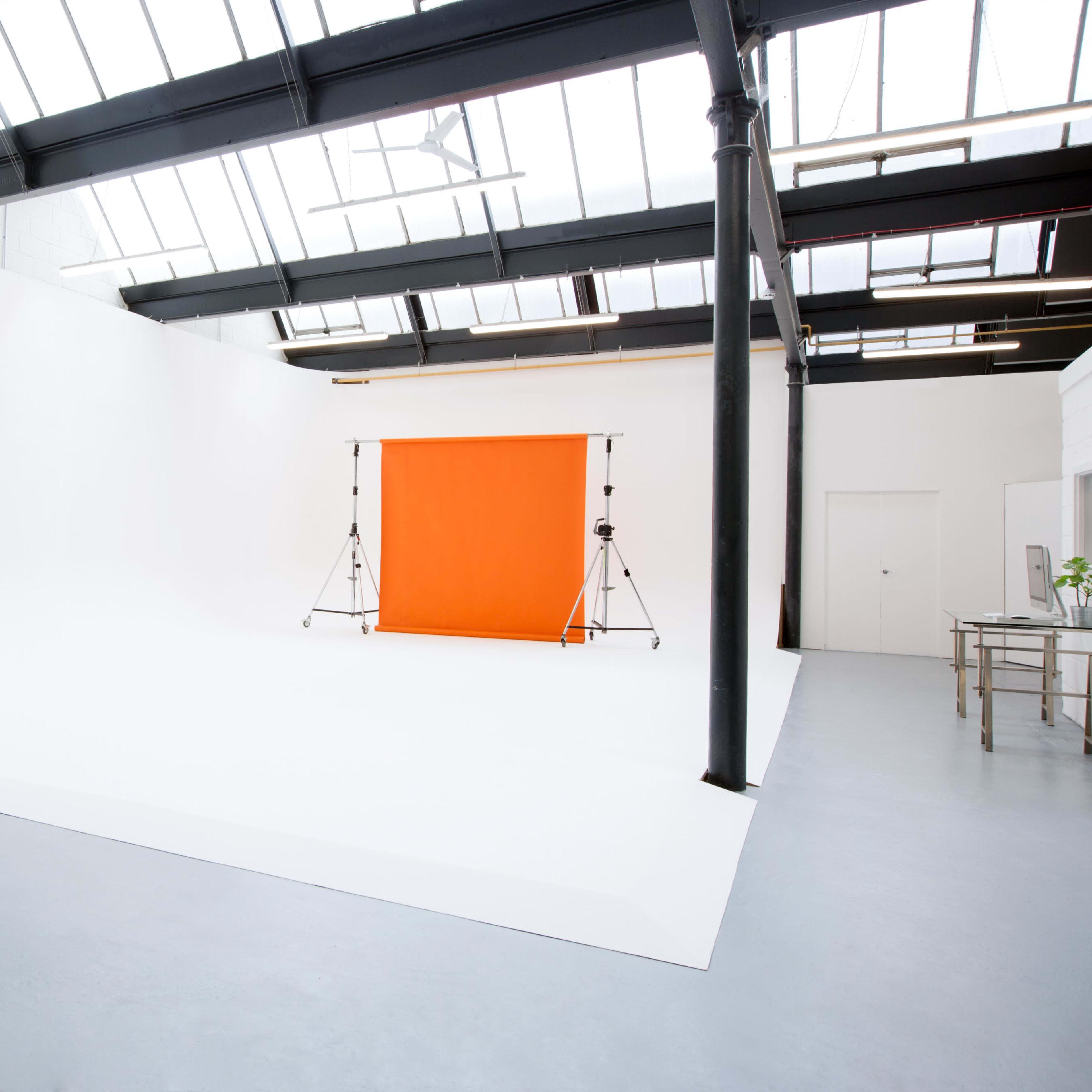 A spacious photography studio with a white backdrop and an orange backdrop set up on stands.