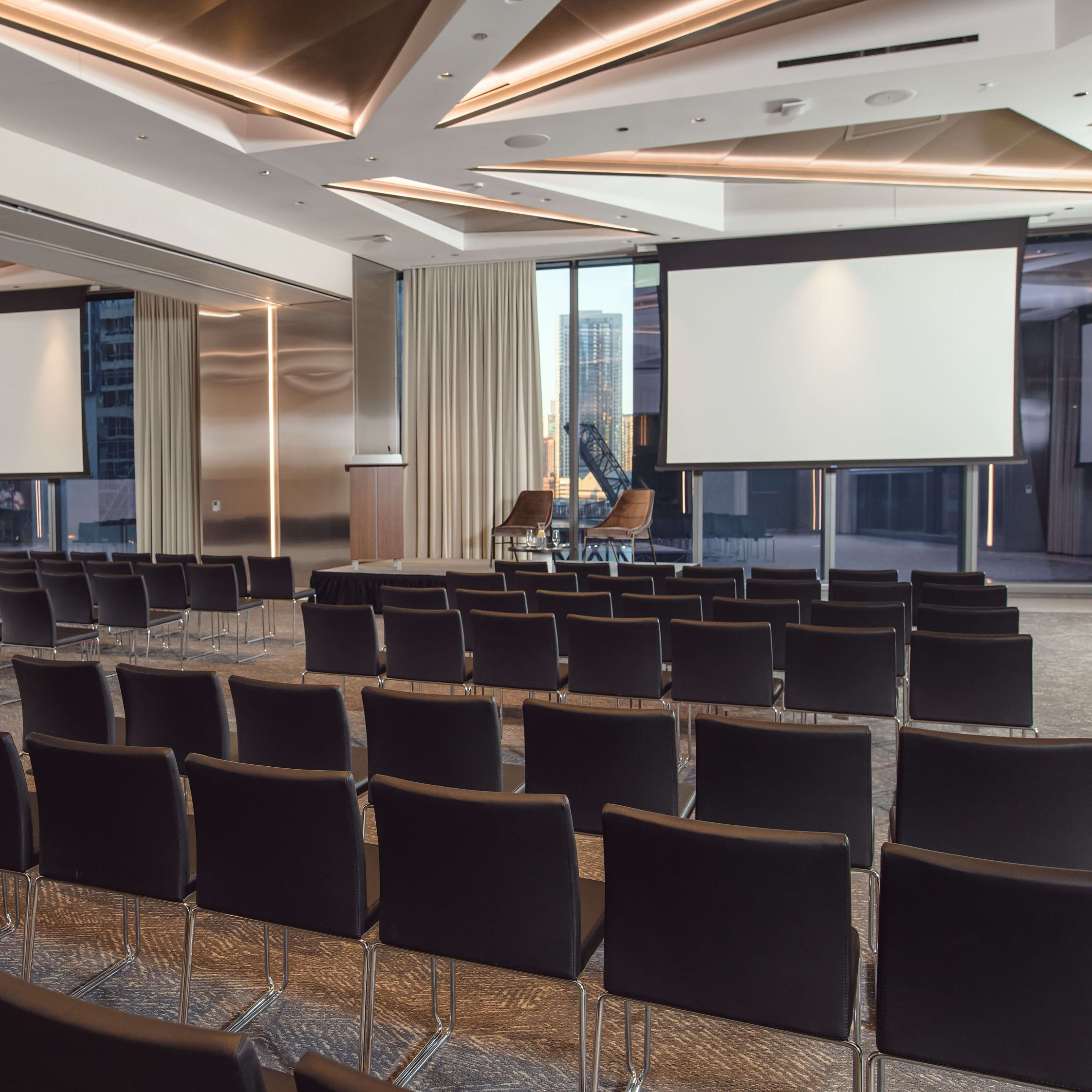 A modern conference room set up with rows of black chairs facing two large screens and a speaker's podium, flanked by large windows overlooking a cityscape.