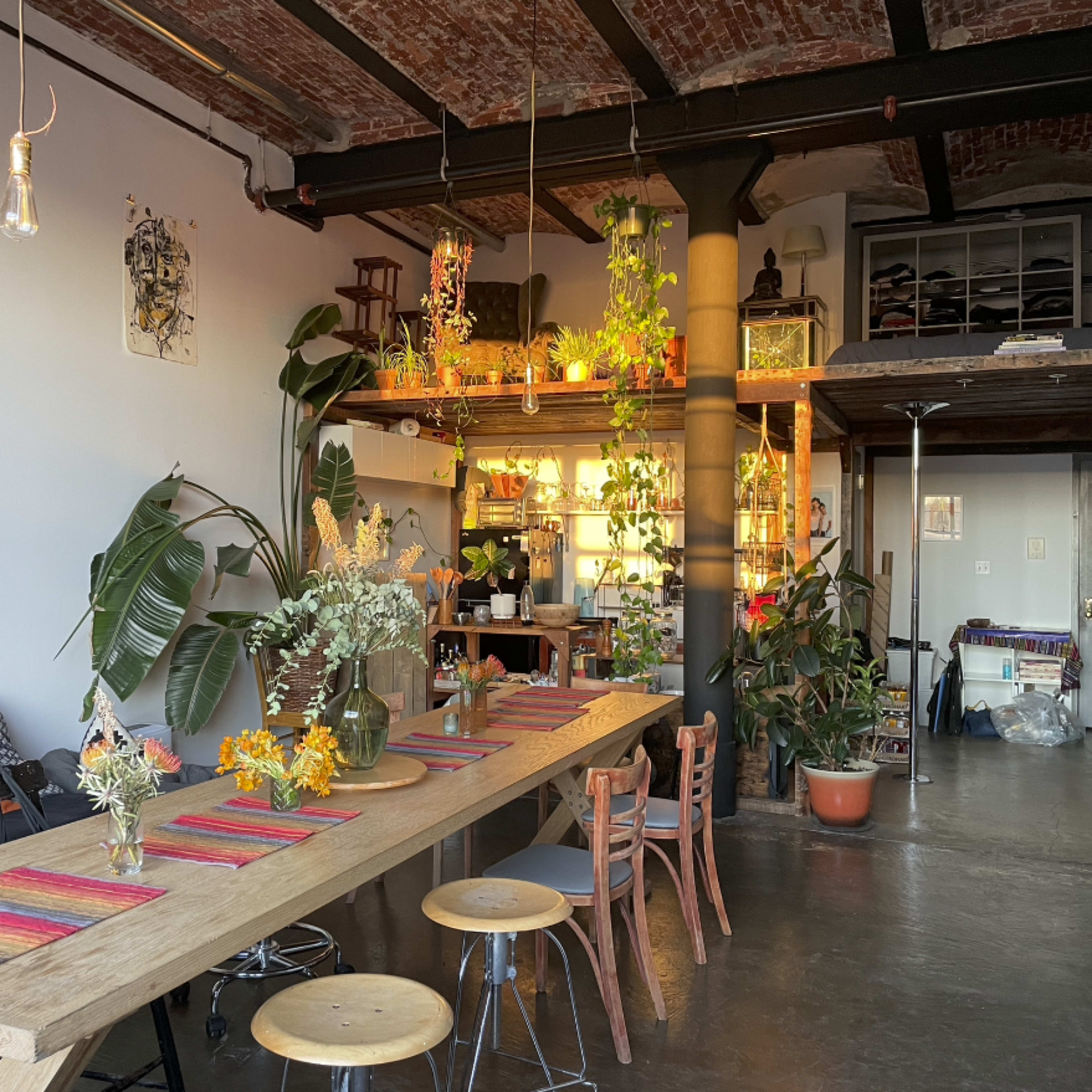 A spacious, well-lit room features a long wooden dining table adorned with colorful placemats, surrounded by plants, chairs, and shelves filled with various items.