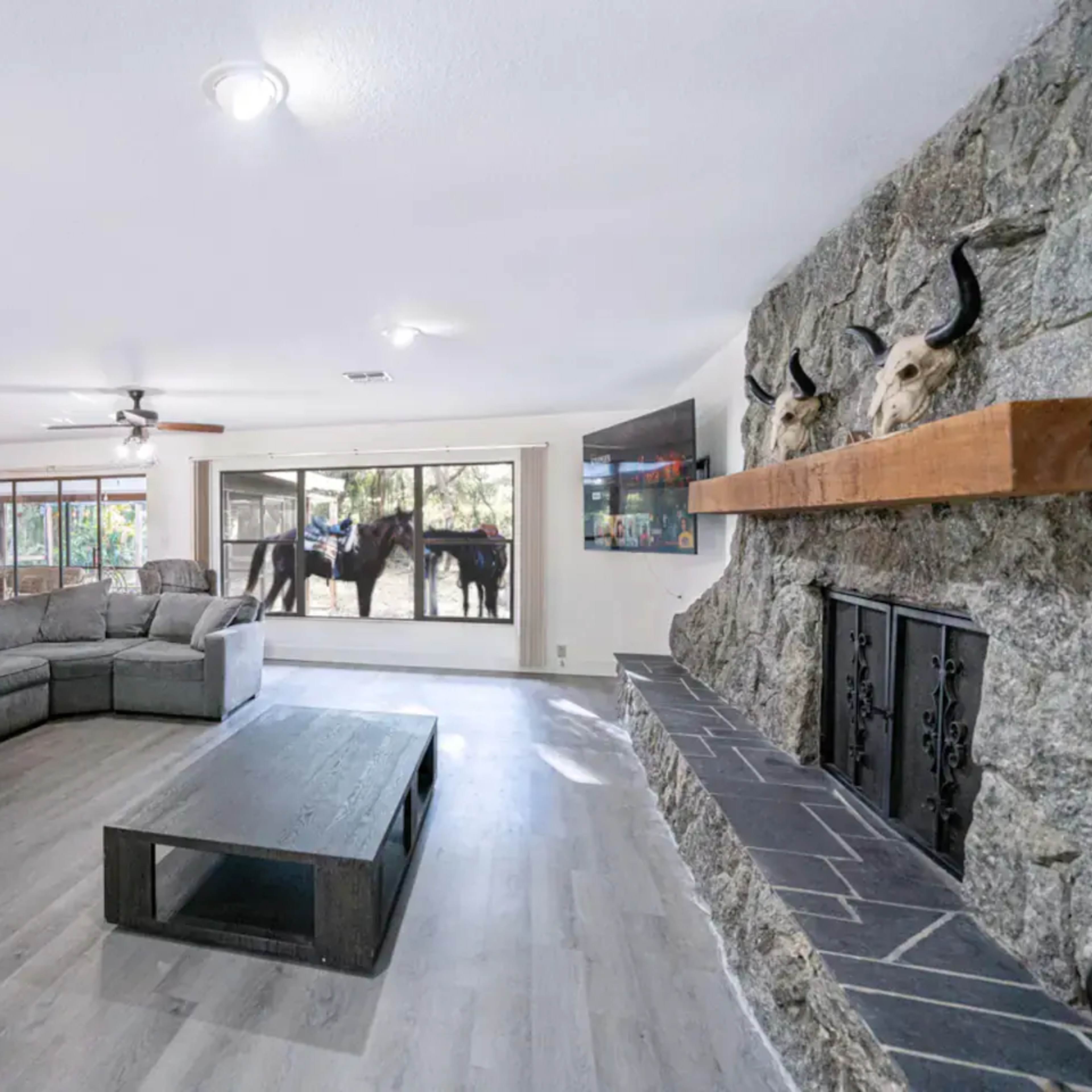 A spacious living room features a large gray sectional sofa, a black coffee table, and a stone fireplace, with large windows showing horses outside.