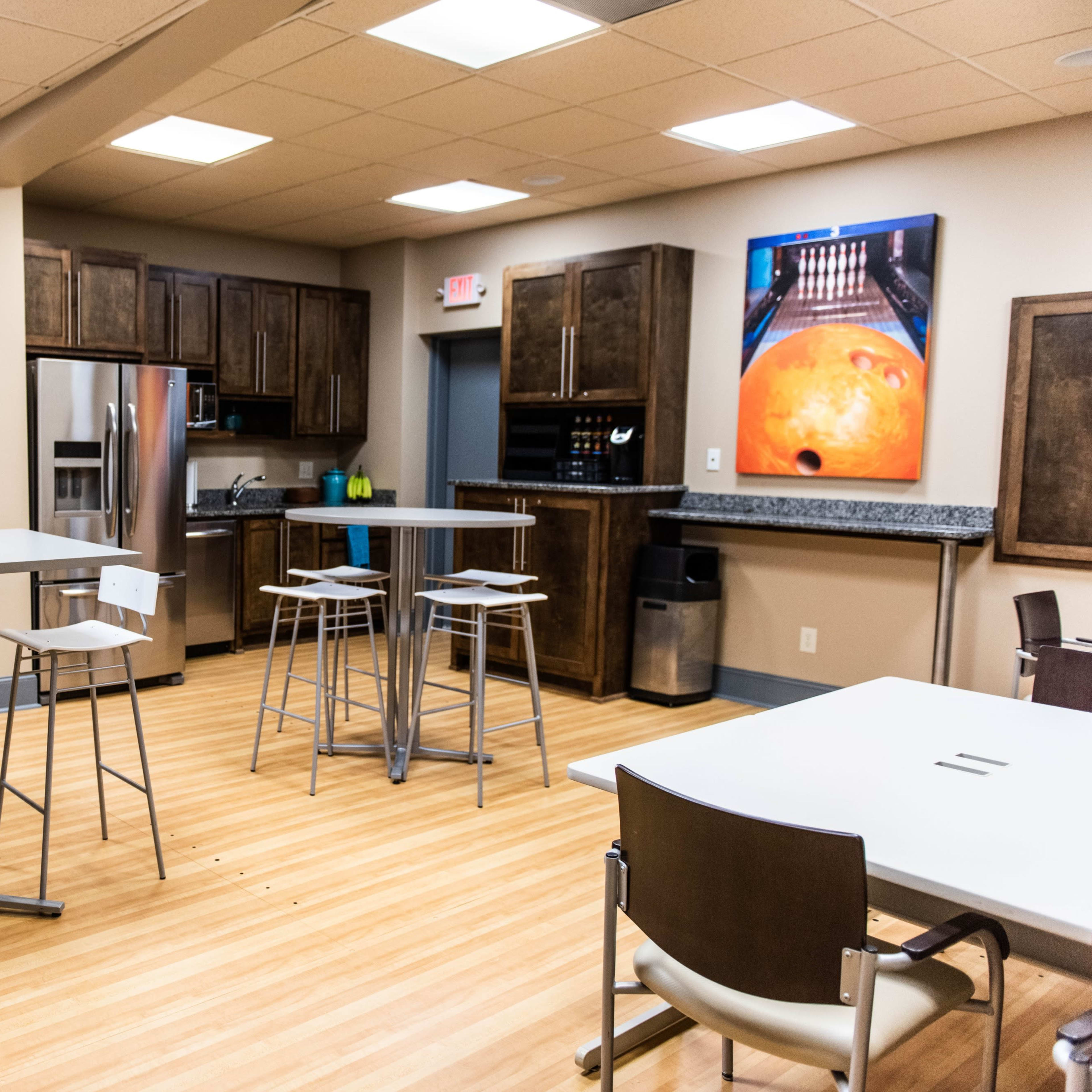 A modern break room featuring several tall tables and stools, a kitchenette with dark wood cabinets, and a television mounted on the wall.