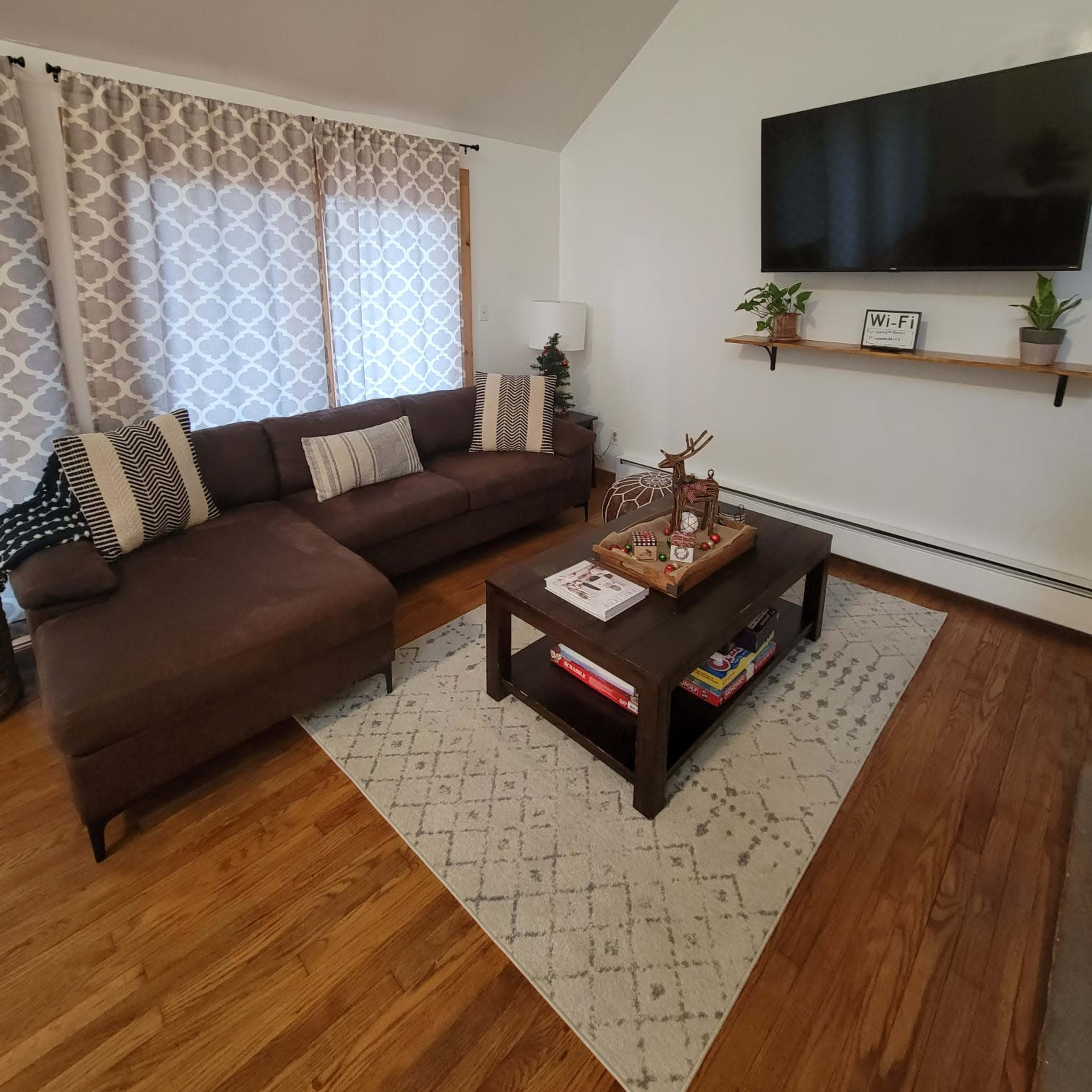The living room features a brown sectional sofa, a wooden coffee table with decorative items, and a wall-mounted television above a shelf with plants.