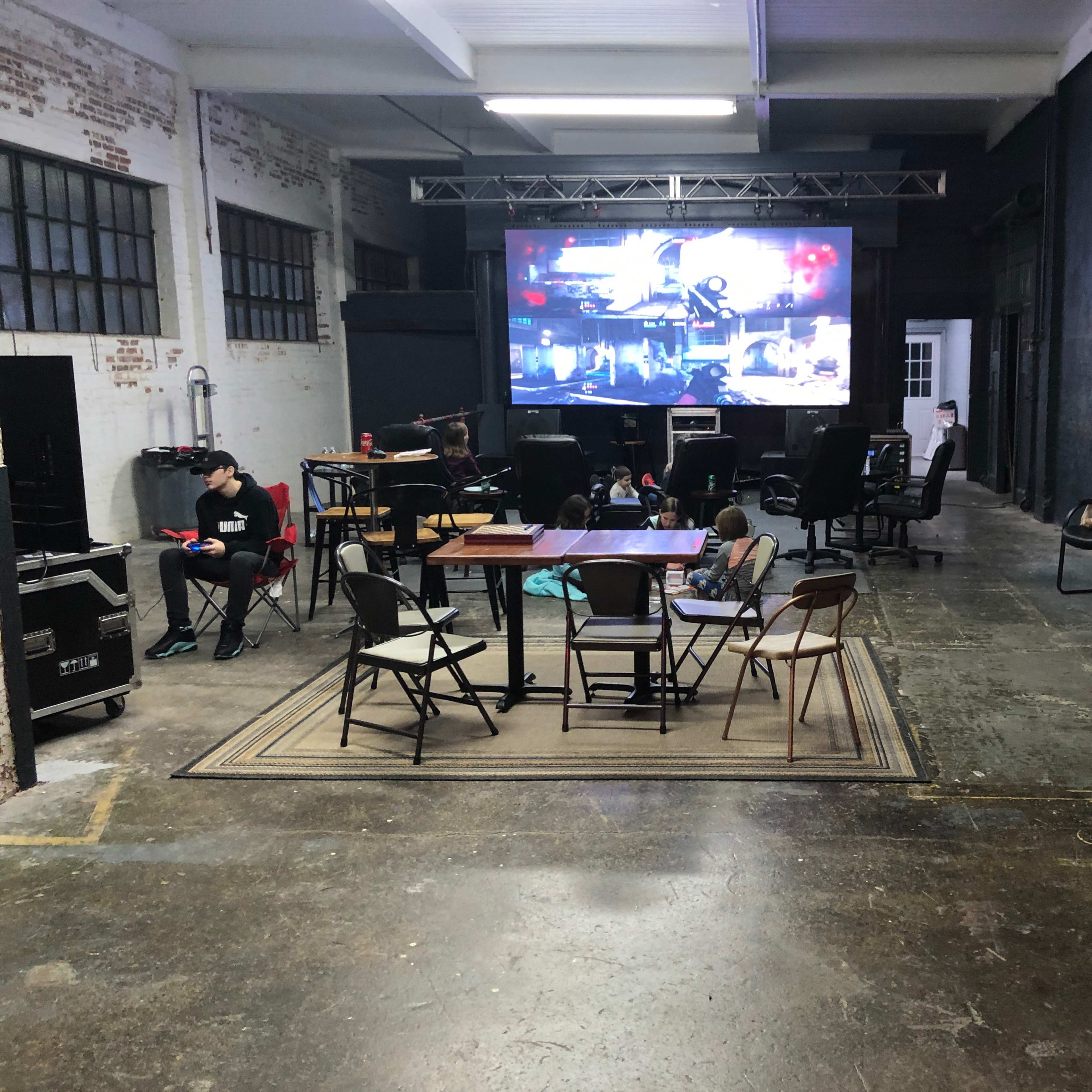 A large screen displays images in an industrial-style room with chairs and tables arranged around a carpet.