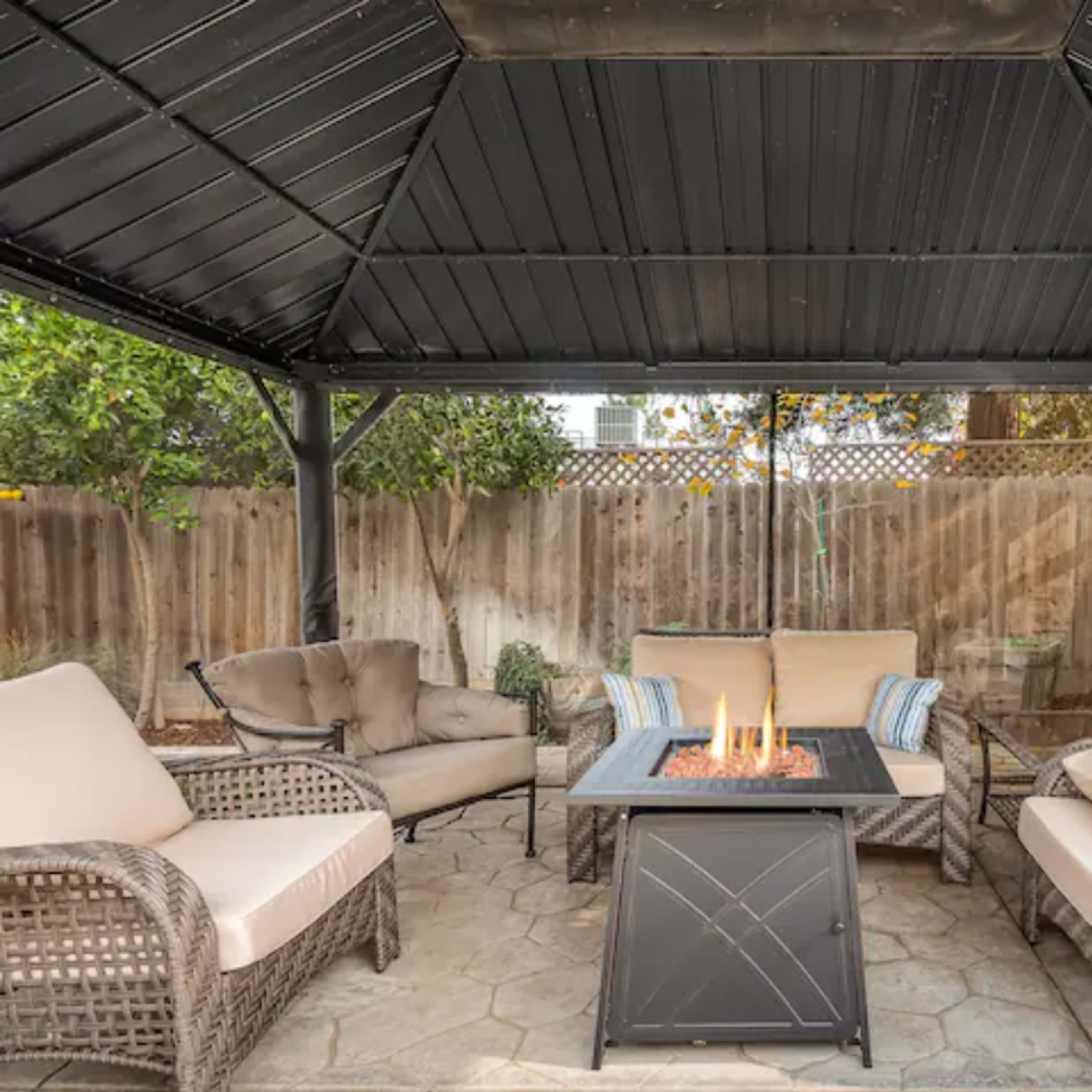 A covered outdoor seating area features a fire pit surrounded by several chairs and a couch on a stone patio.